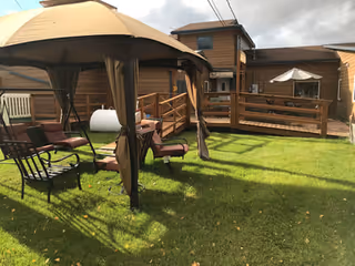 Outdoor grassy area with a brown gazebo canopy covering several chairs and a small table. The area is adjacent to a wooden building with a ramp and railing, and there is a white umbrella on a patio in the background under a cloudy sky.