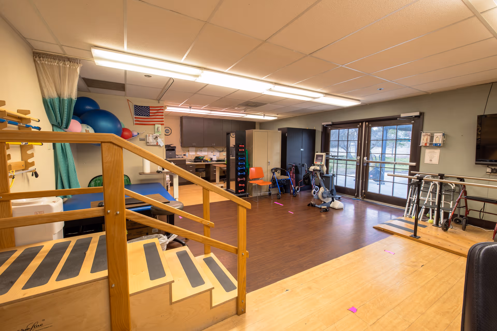 A rehabilitation or physical therapy room with wooden stairs and handrails for practice, exercise equipment including a stationary bike, walkers, and therapy balls. The room has large windows and double doors leading outside, with cabinets and an American flag on the wall.