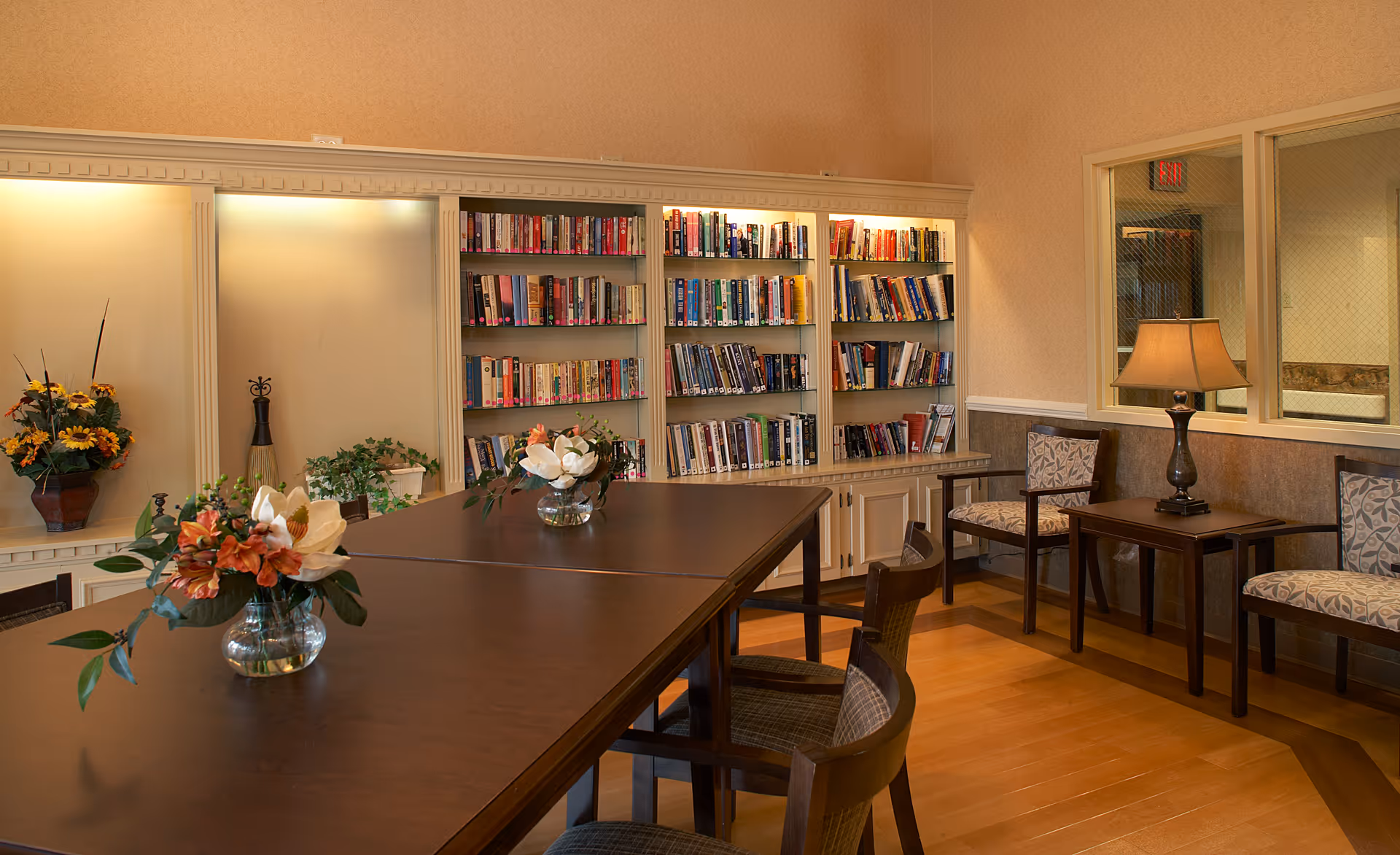 A cozy interior room with a large wooden table surrounded by chairs. The back wall features built-in bookshelves filled with books and decorative plants. There are additional chairs and a side table with a lamp along the right wall. The room has warm lighting and a wooden floor.