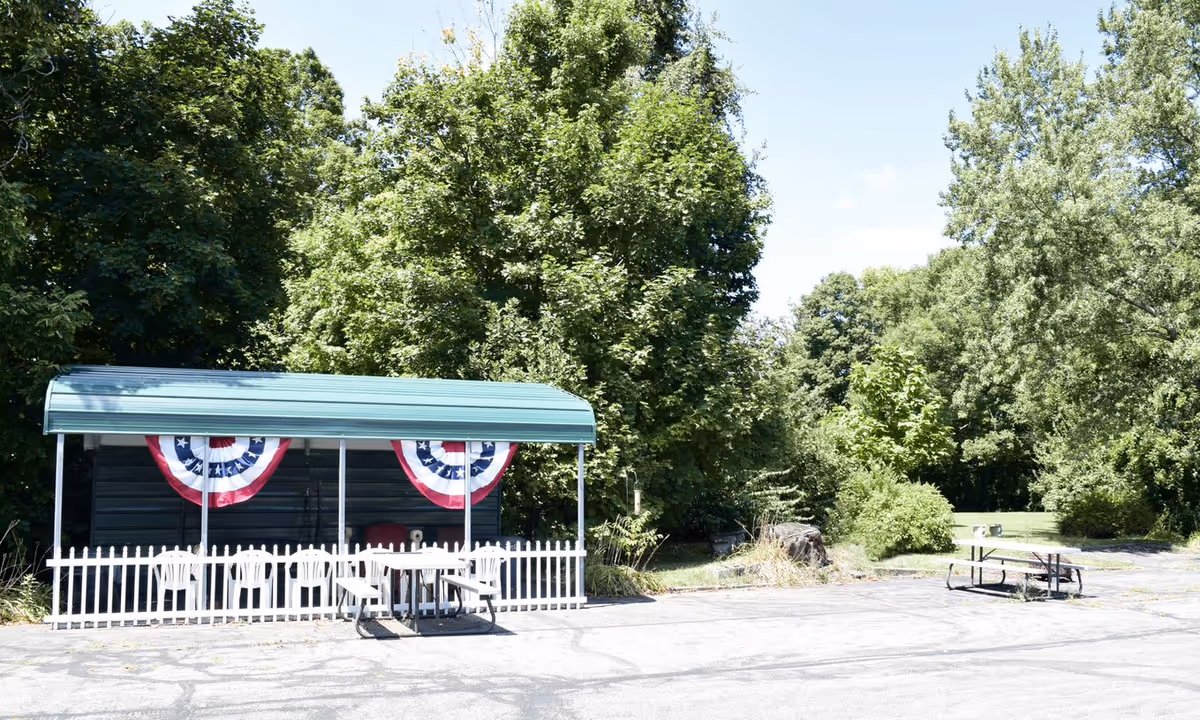 A covered outdoor seating area with white chairs and patriotic bunting among trees.
