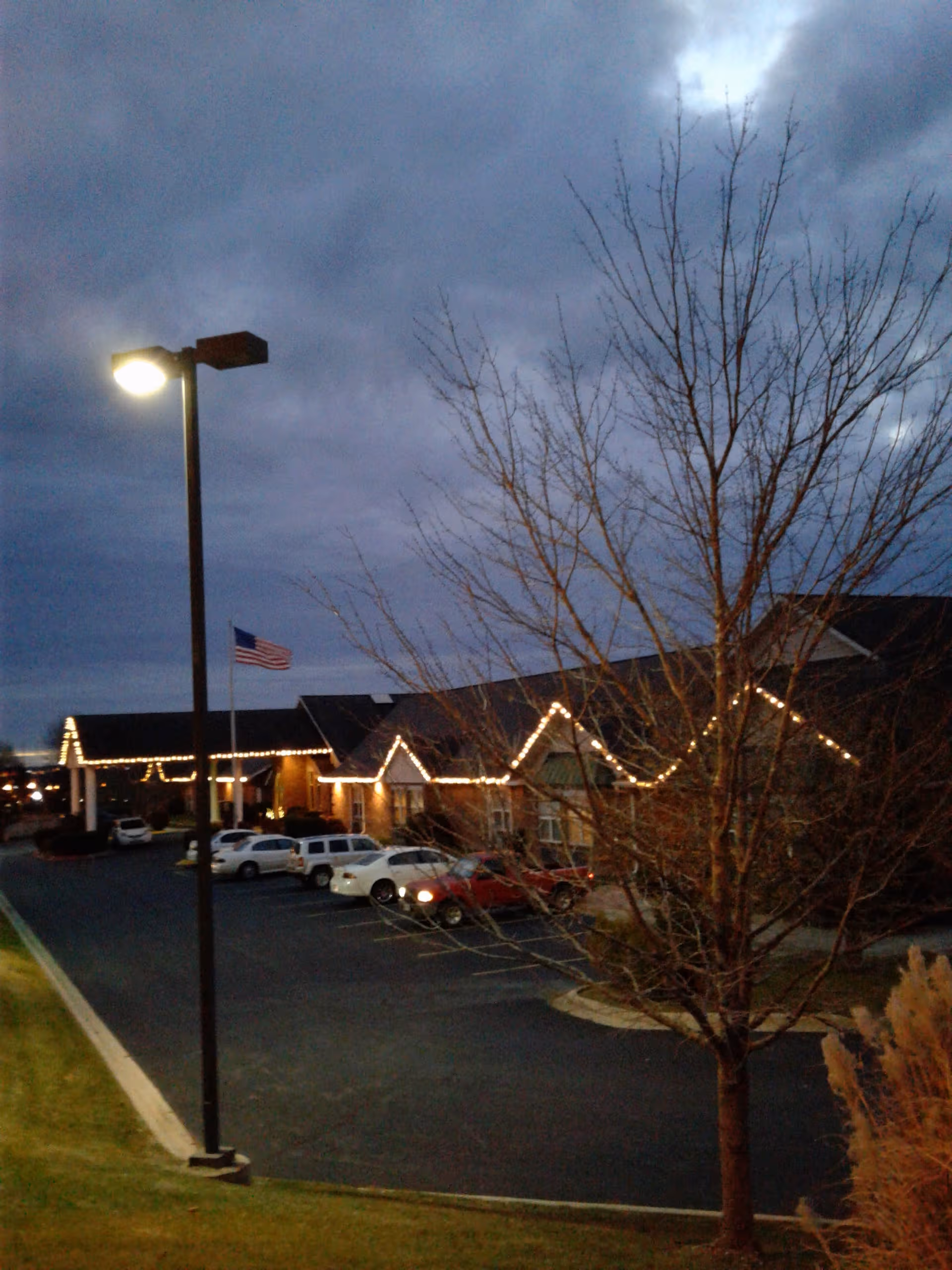 Evening exterior of a senior living facility with string lights, parked cars, and bare trees.