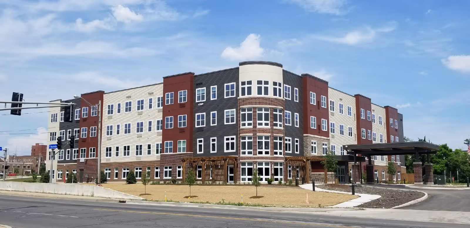 Exterior view of a modern four-story senior living facility building with multiple windows, a covered entrance, and a landscaped area with small trees and grass. The sky is partly cloudy.