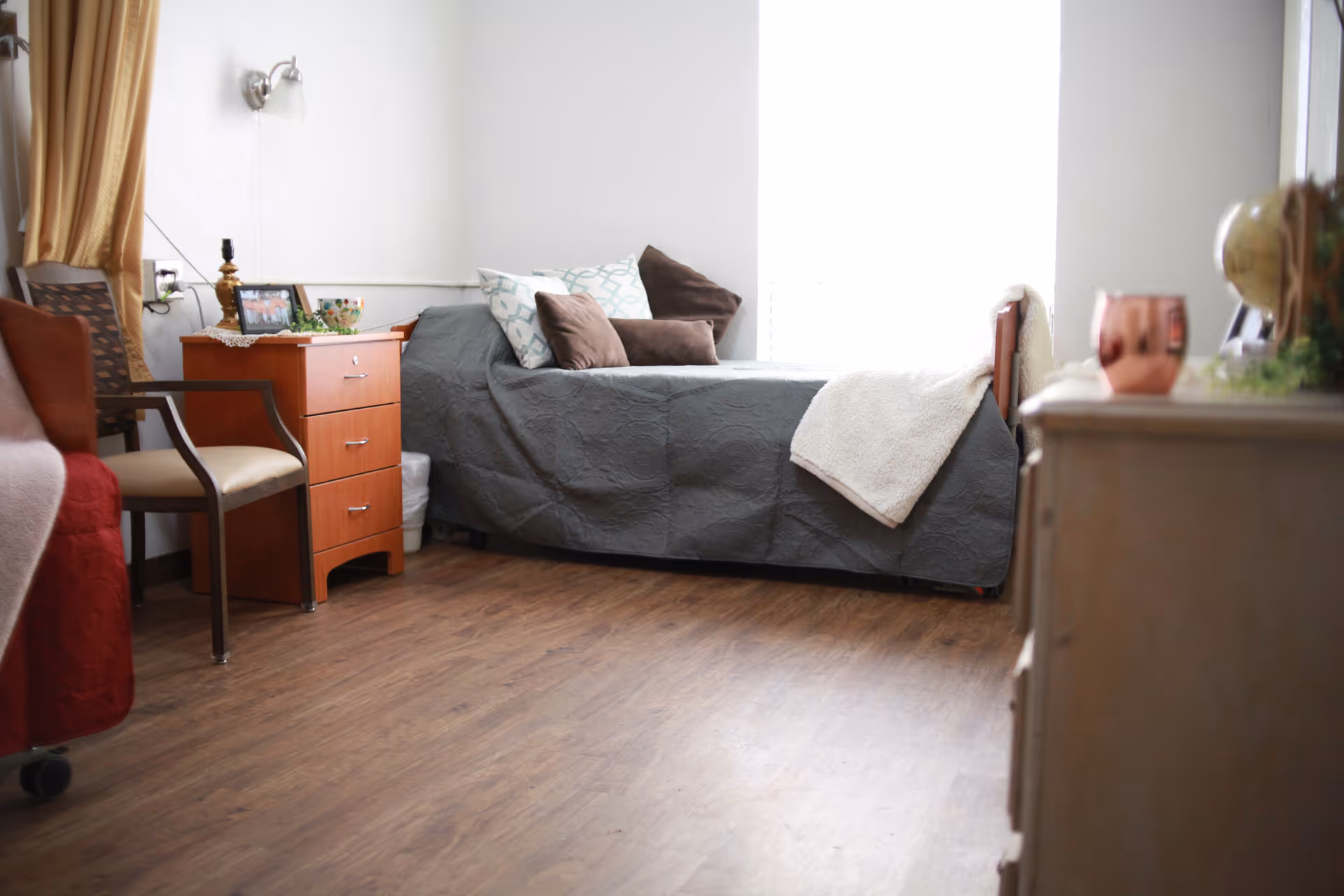 A tidy senior living bedroom with a gray bedspread, dresser, chair, and window.