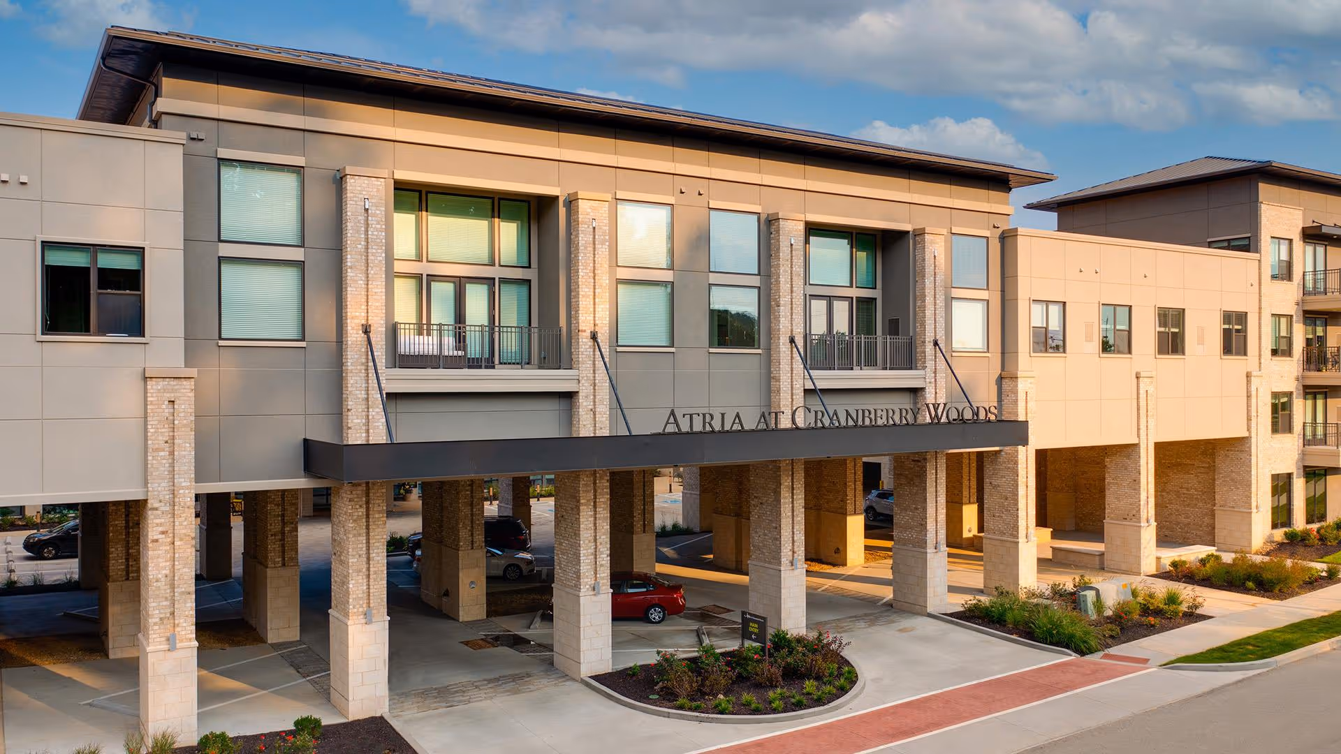 Exterior view of a modern senior living facility building with multiple floors, large windows, and a covered entrance supported by brick pillars. The building is surrounded by landscaped greenery and a paved driveway.