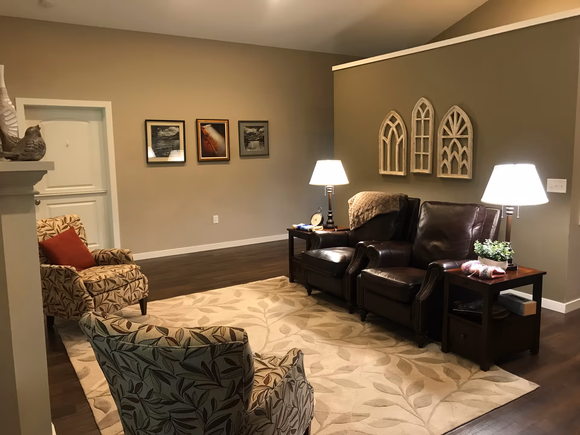 A cozy living room area with two patterned armchairs and two dark leather armchairs arranged around a beige rug with leaf patterns. There are two side tables with lamps, a small plant, and decorative items. The walls are painted in neutral tones with three framed pictures on one wall and three decorative window-shaped wall hangings on another.