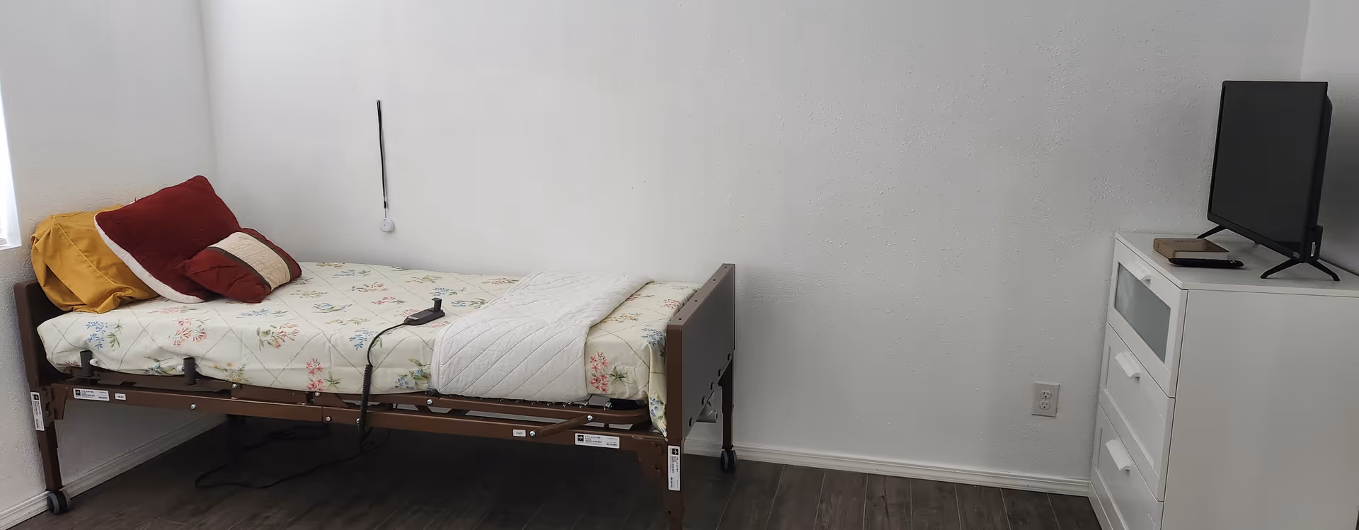 A hospital-style single bed with floral sheets, pillows, and a white dresser with a TV.
