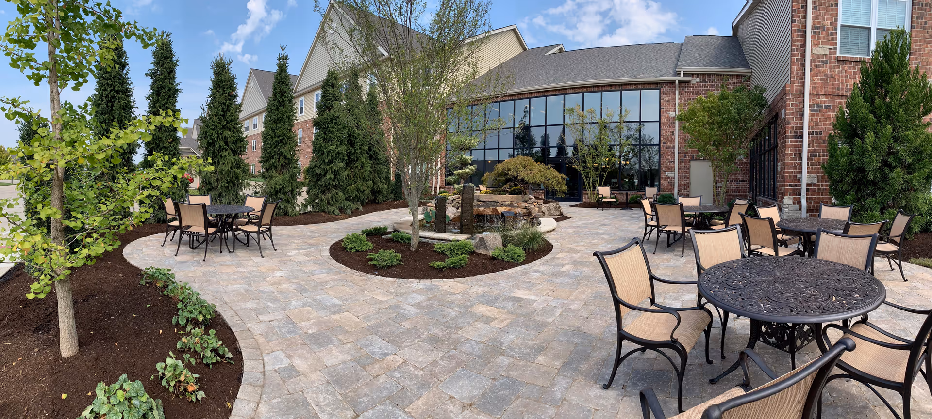 Outdoor courtyard patio with paved walkways, metal tables and chairs, landscaped plantings and the building's brick facade with large windows.
