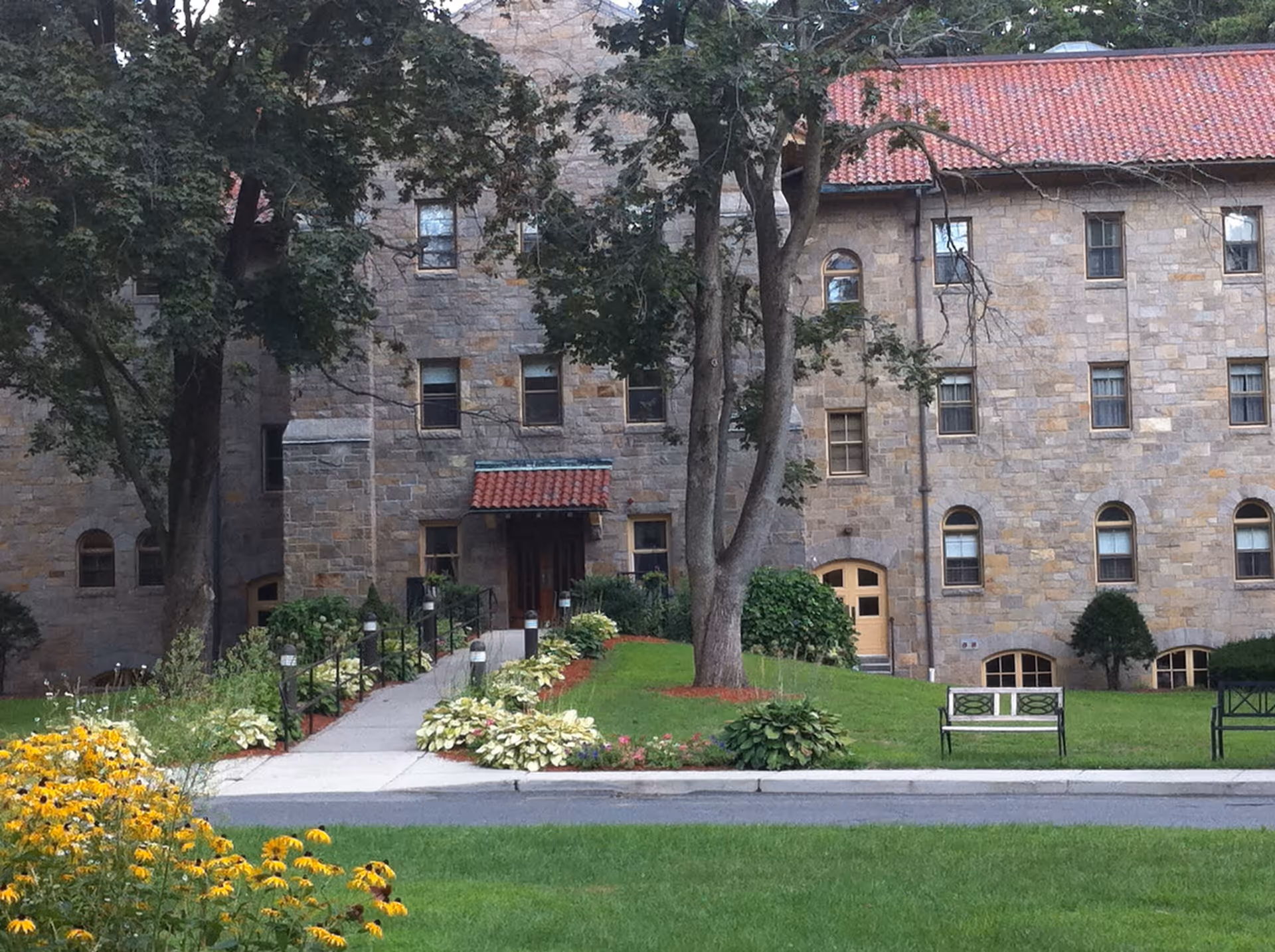 Stone building with multiple windows and a red tiled roof, surrounded by green grass, trees, flower beds, and benches along a paved walkway leading to an entrance with a small red awning.