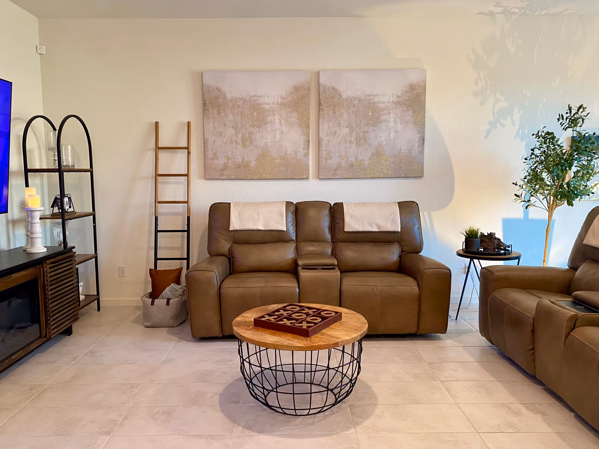 A cozy living room with two brown leather recliners and a matching loveseat, a round wooden coffee table with a tic-tac-toe game on top, a black metal and wood shelving unit with decorative items, two abstract paintings on the wall, a small side table with a plant, and a potted tree in the corner.