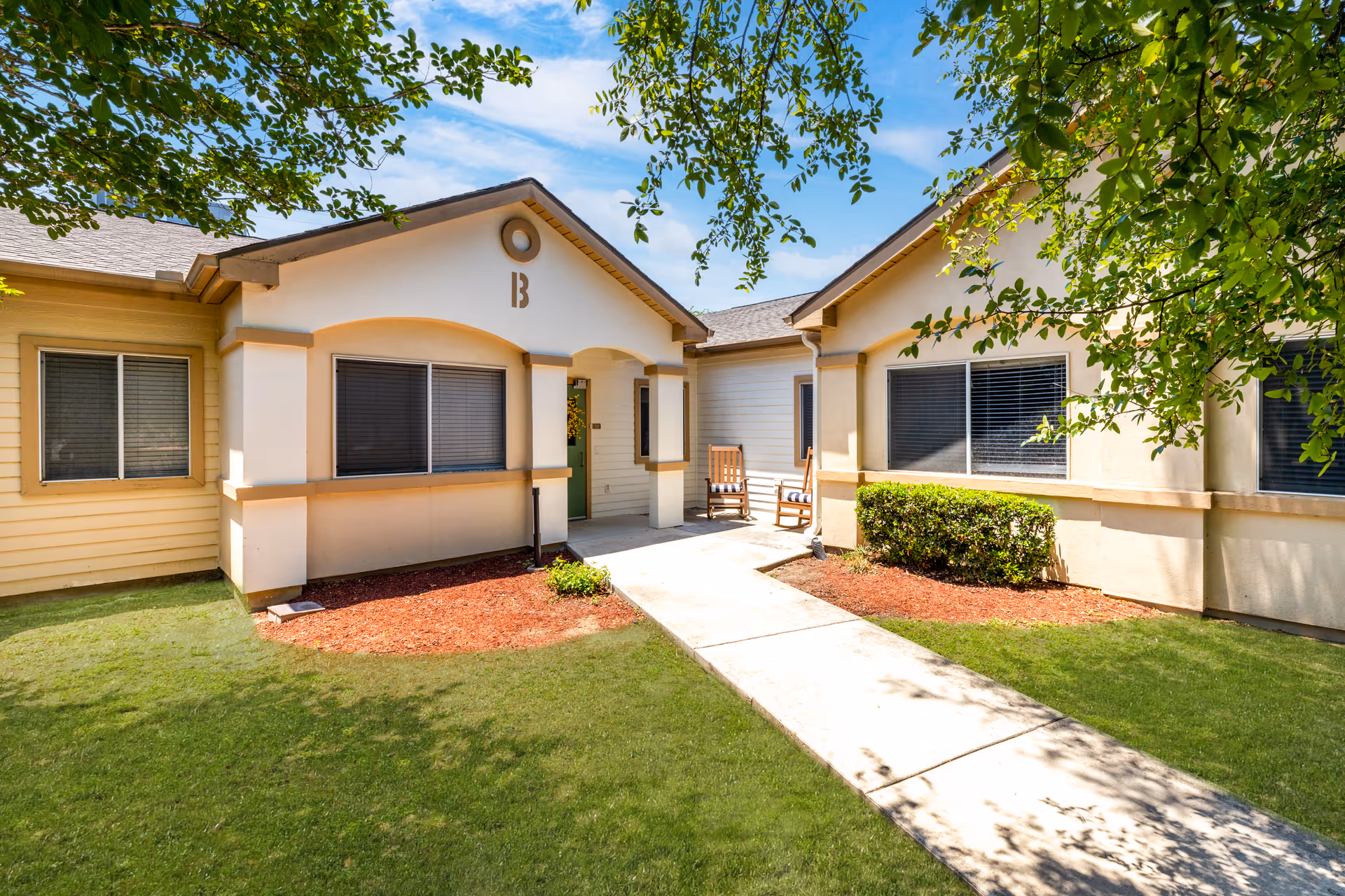 Exterior of a single-story senior living building with a walkway, chairs, and shaded entrance.