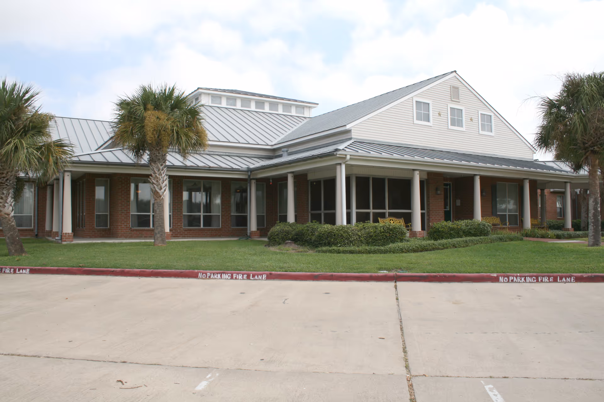 Exterior view of Villa of Corpus Christi South, a brick senior living facility with palm trees.