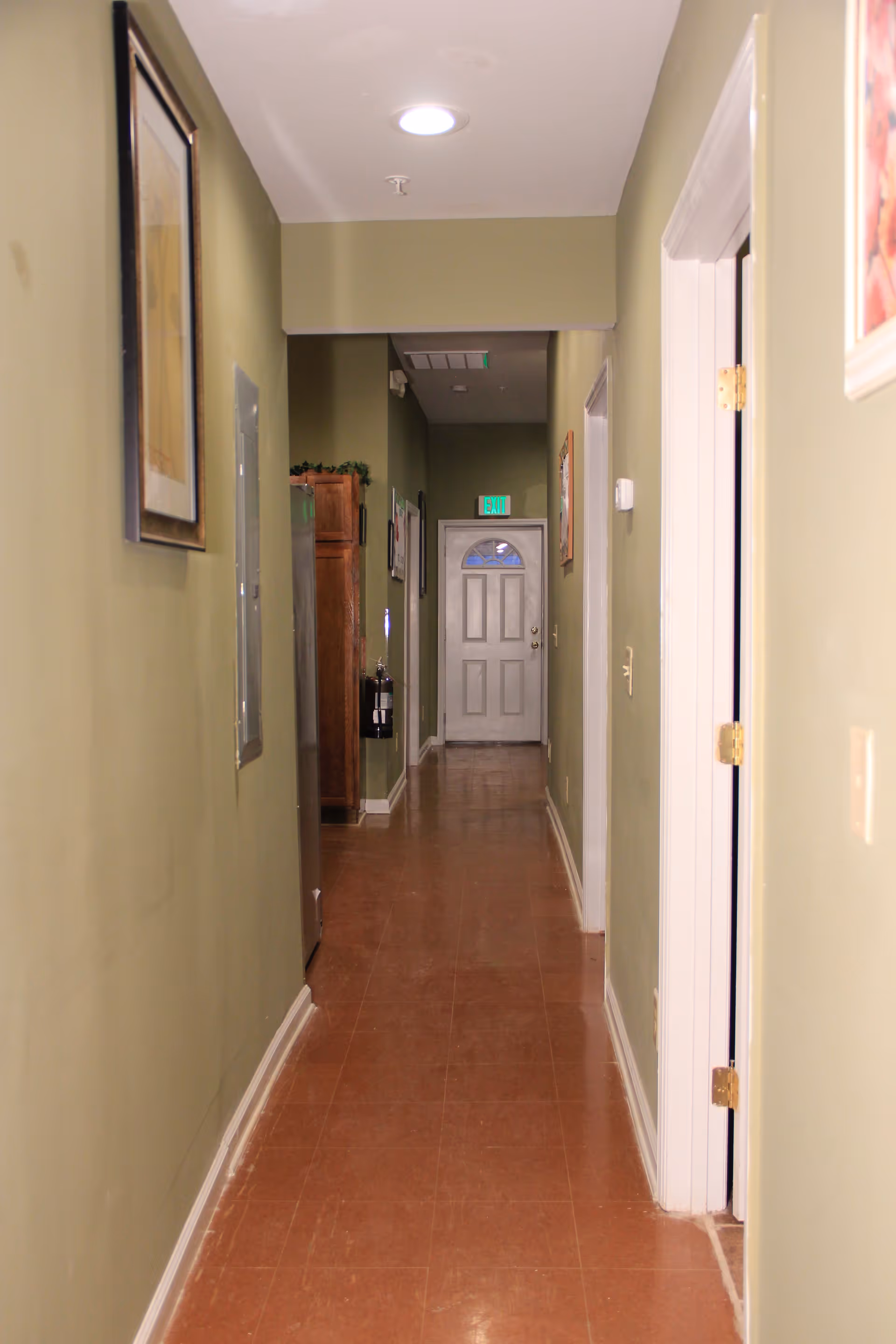 Long narrow interior hallway with green walls, framed pictures, and a white door with an exit sign at the far end.
