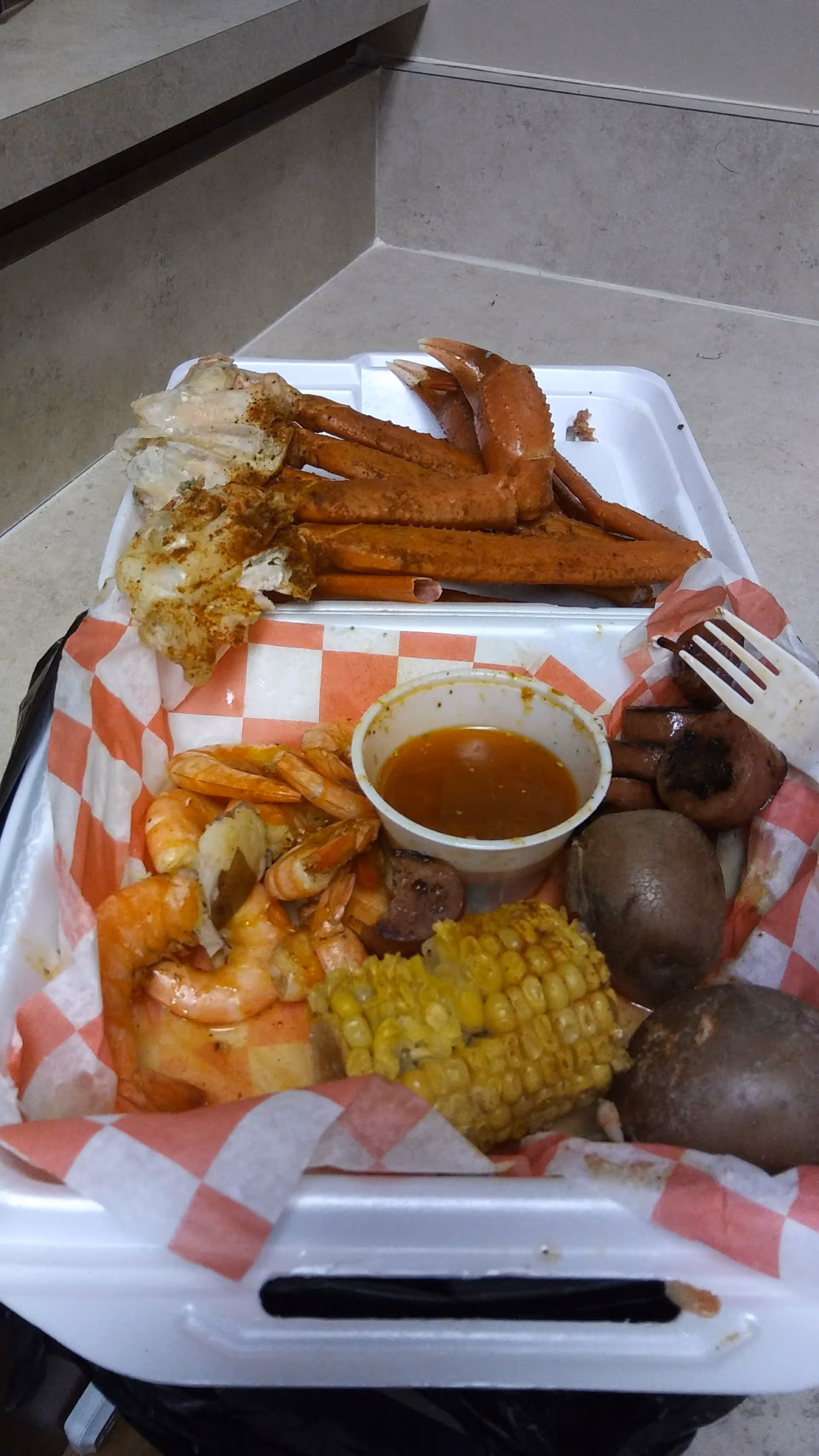 A takeout-style tray with crab legs, shrimp, corn, potatoes, and sauce.