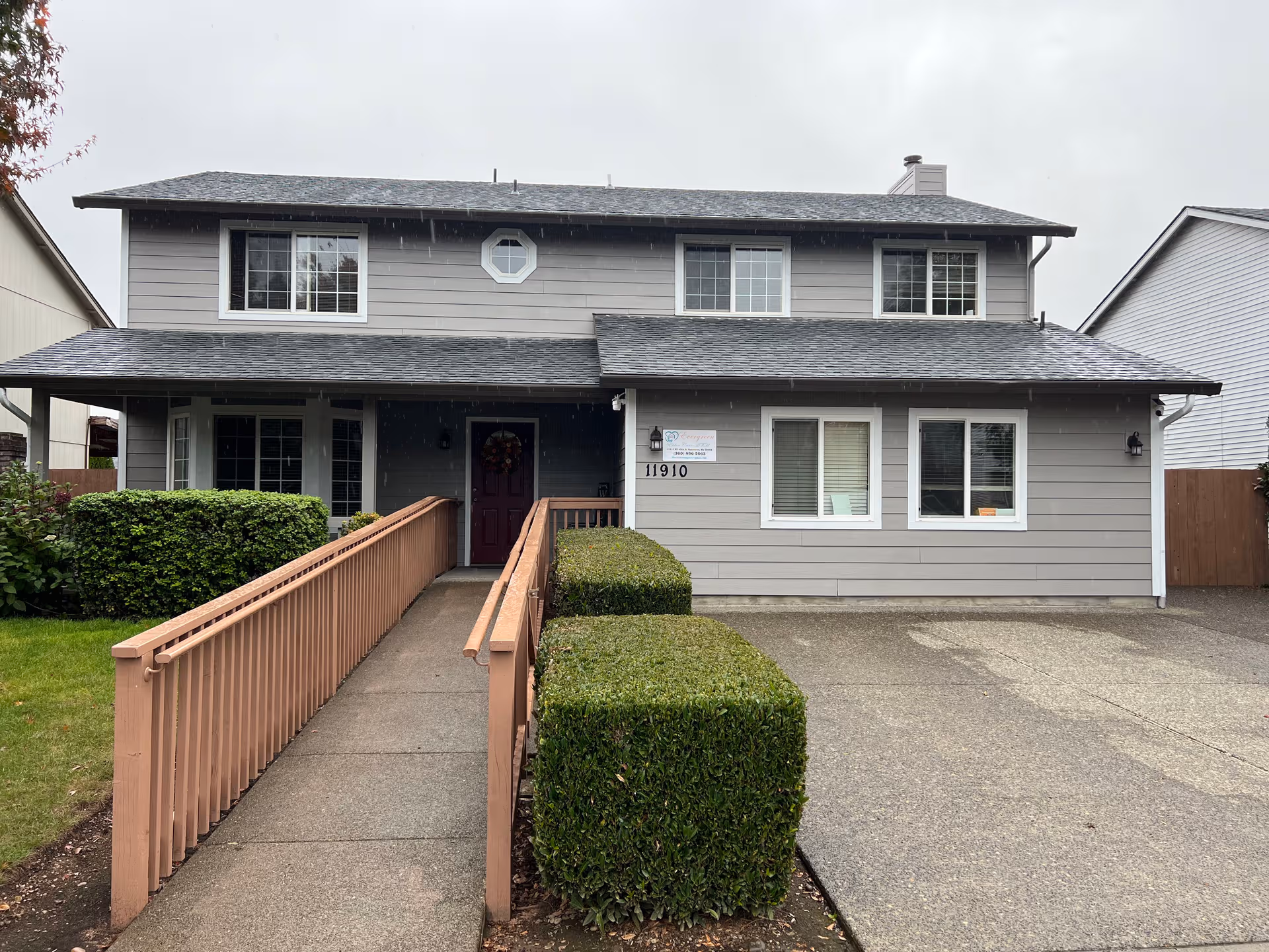 Two-story gray adult family home with ramped front walkway and trimmed hedges.