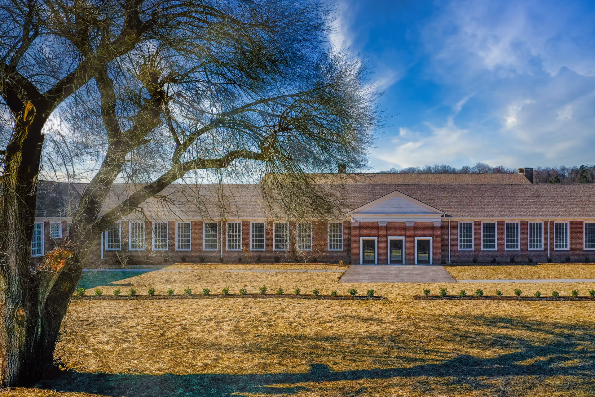 Front exterior of a long brick senior living building with a central entrance and a large leafless tree in the foreground.