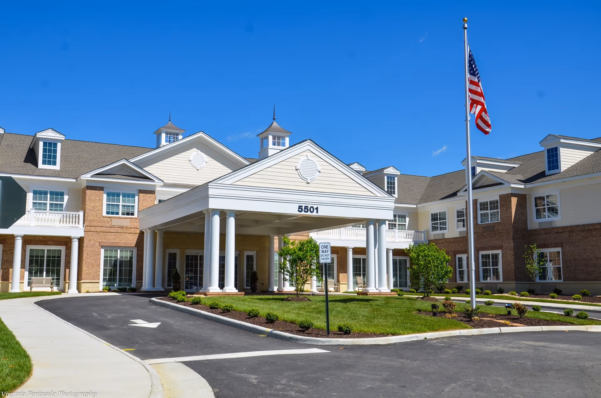 Front exterior view of a senior living facility named Edgeworth Park at New Town, featuring a large covered entrance with white columns, a manicured lawn, an American flag on a flagpole, and a clear blue sky.