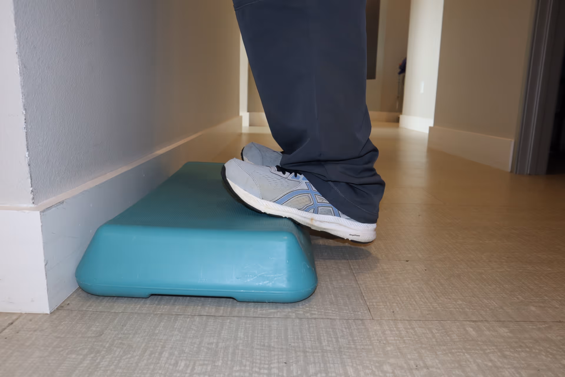 Lower legs in sneakers stand with one foot on a turquoise step platform in a hallway.