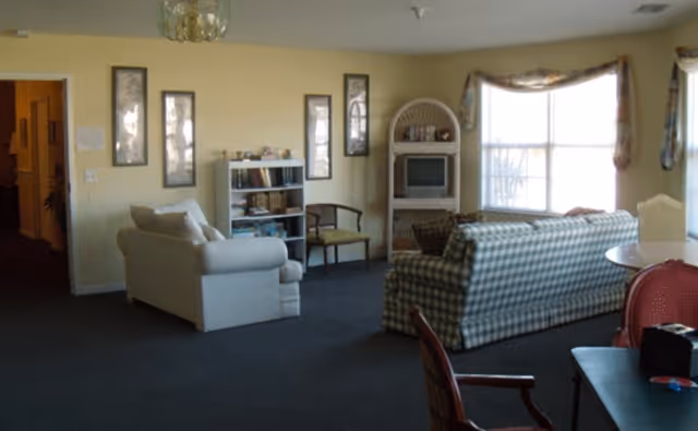 A communal living room with sofas, a TV, a bookshelf, and large windows.