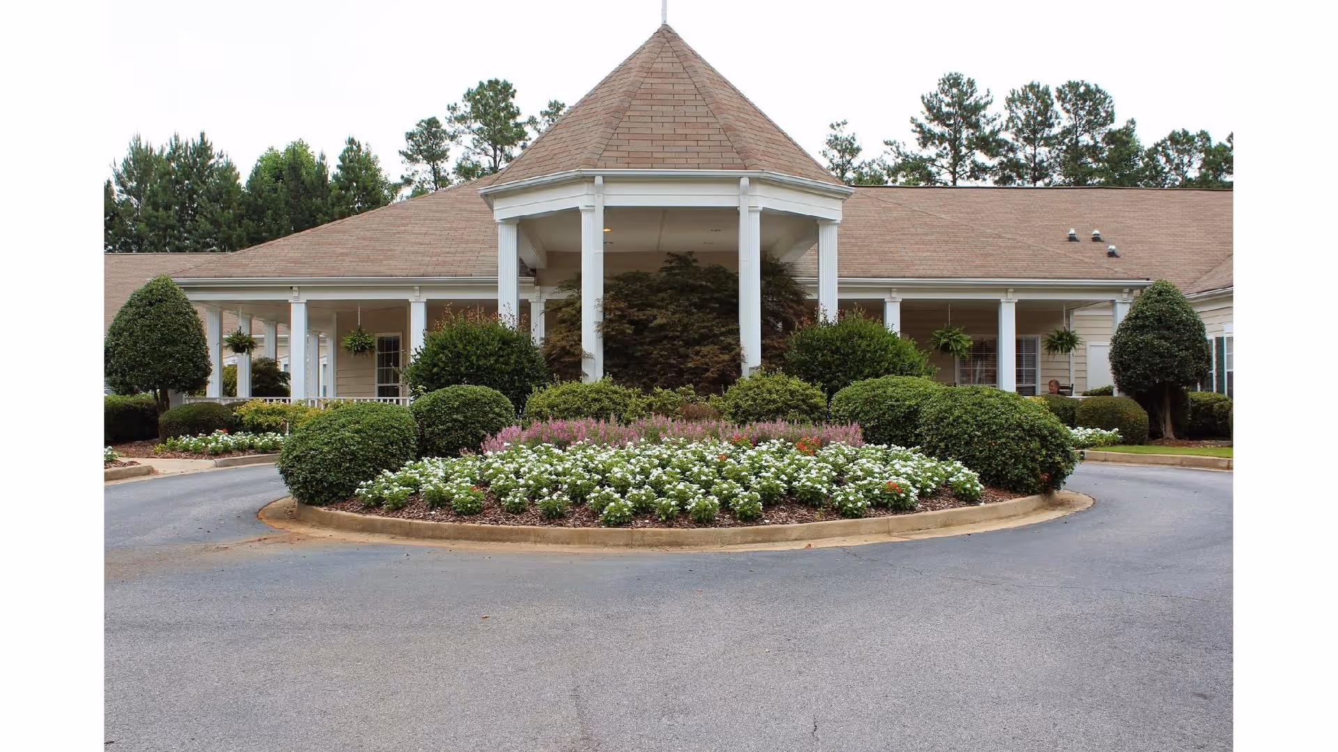 Front exterior view of Oaks at Cedar Shoals featuring a circular driveway with a landscaped roundabout filled with bushes and flowers, a covered entrance supported by white columns, and a building with a beige roof surrounded by trees.