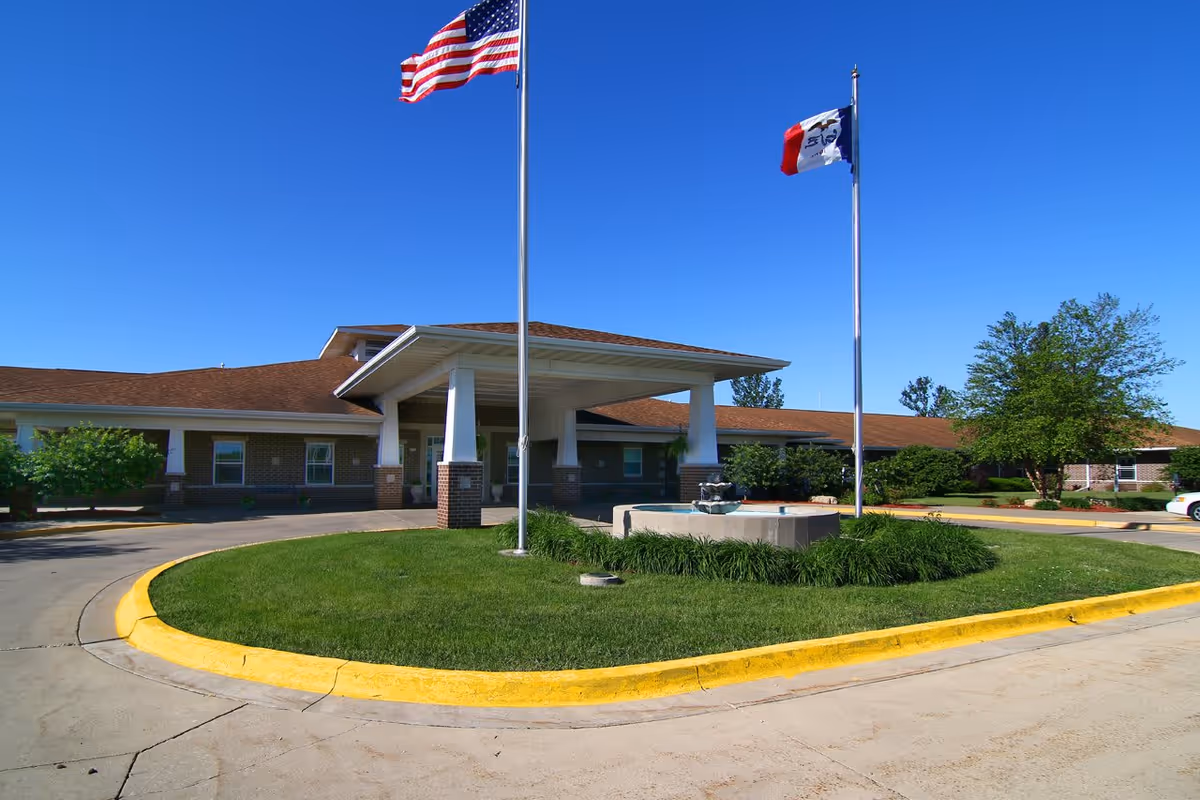 Exterior view of Prairie Hills at Independence facility showing a single-story brick building with a covered entrance, two flagpoles with the American flag and another flag, a circular driveway with a grassy island and a small fountain, under a clear blue sky.