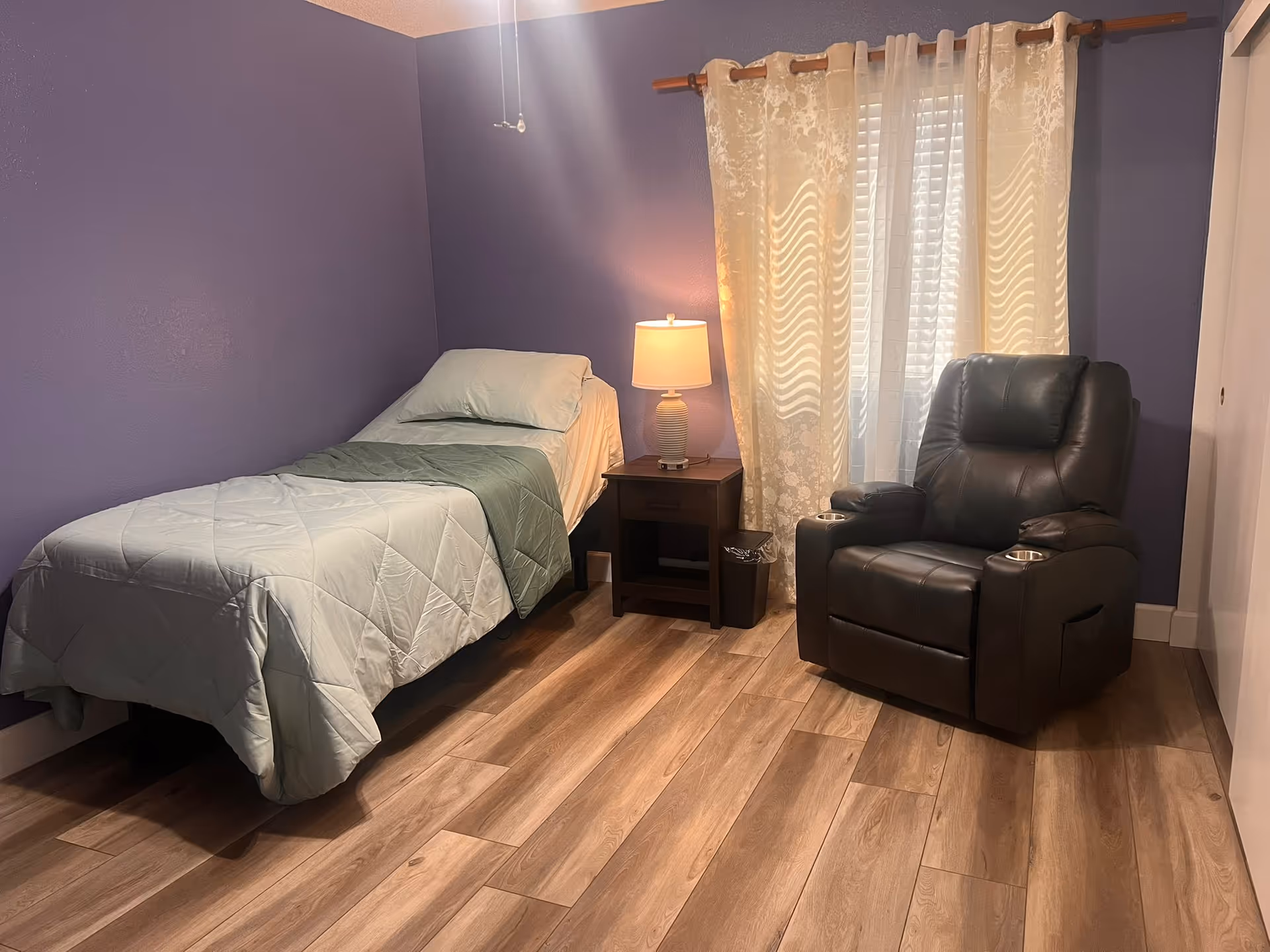 Purple-walled bedroom featuring a bed, nightstand, lamp, window curtains, and a recliner.