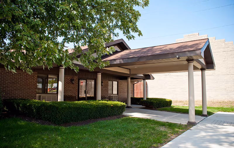 Covered entrance to a brick building with columns supporting a brown roof, surrounded by green bushes and a tree with green leaves under a clear blue sky.