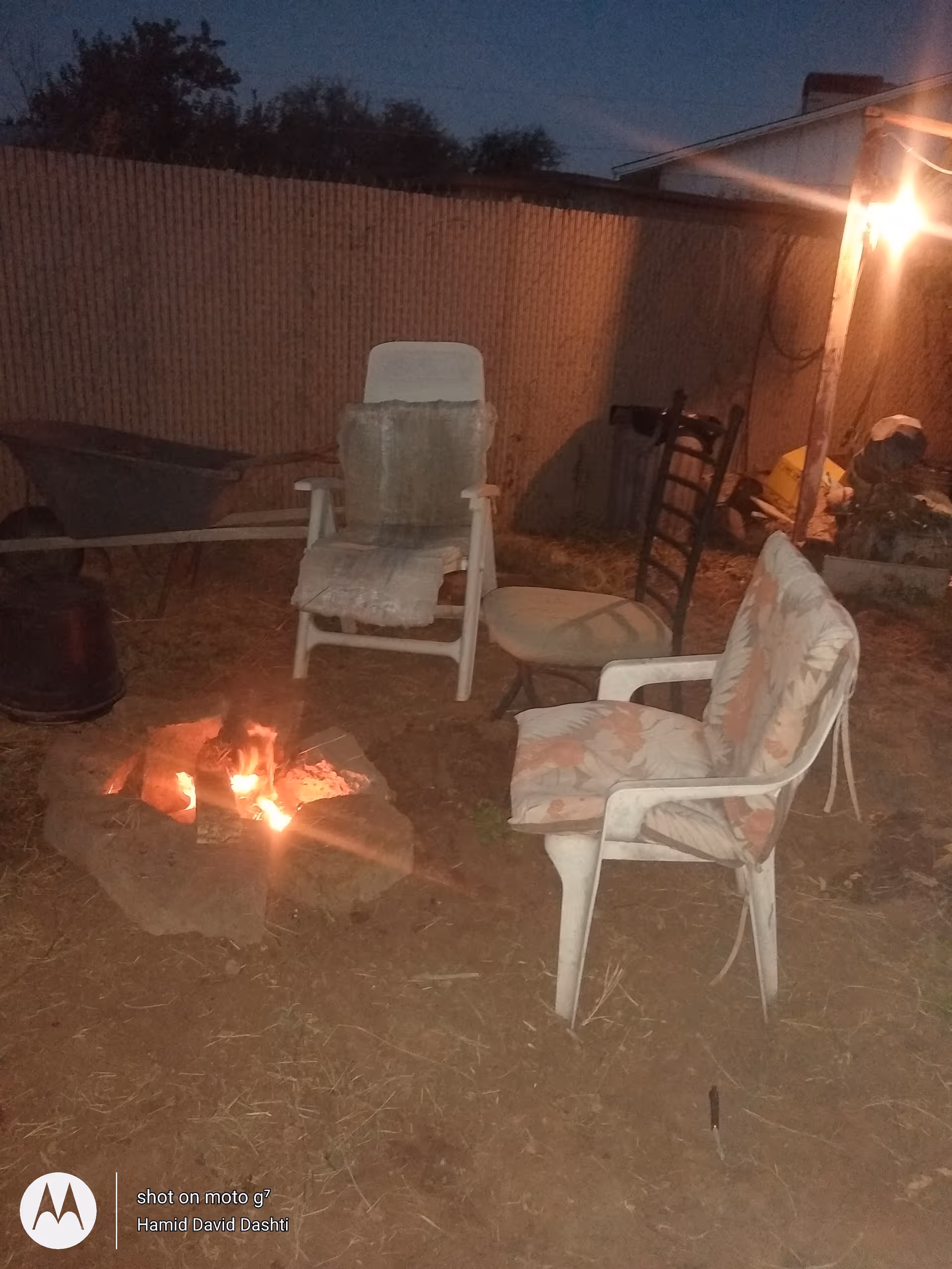 Outdoor evening scene with a lit fire pit and mismatched chairs around it.
