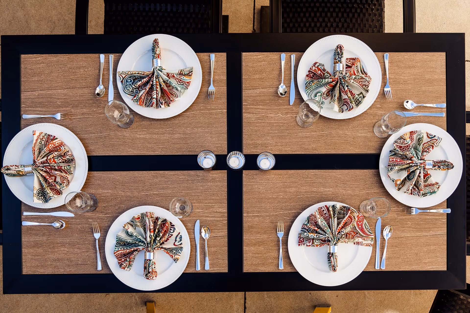 Overhead view of a dining table set for six people with white plates, colorful patterned napkins folded in a fan shape with napkin rings, silverware, and clear drinking glasses.