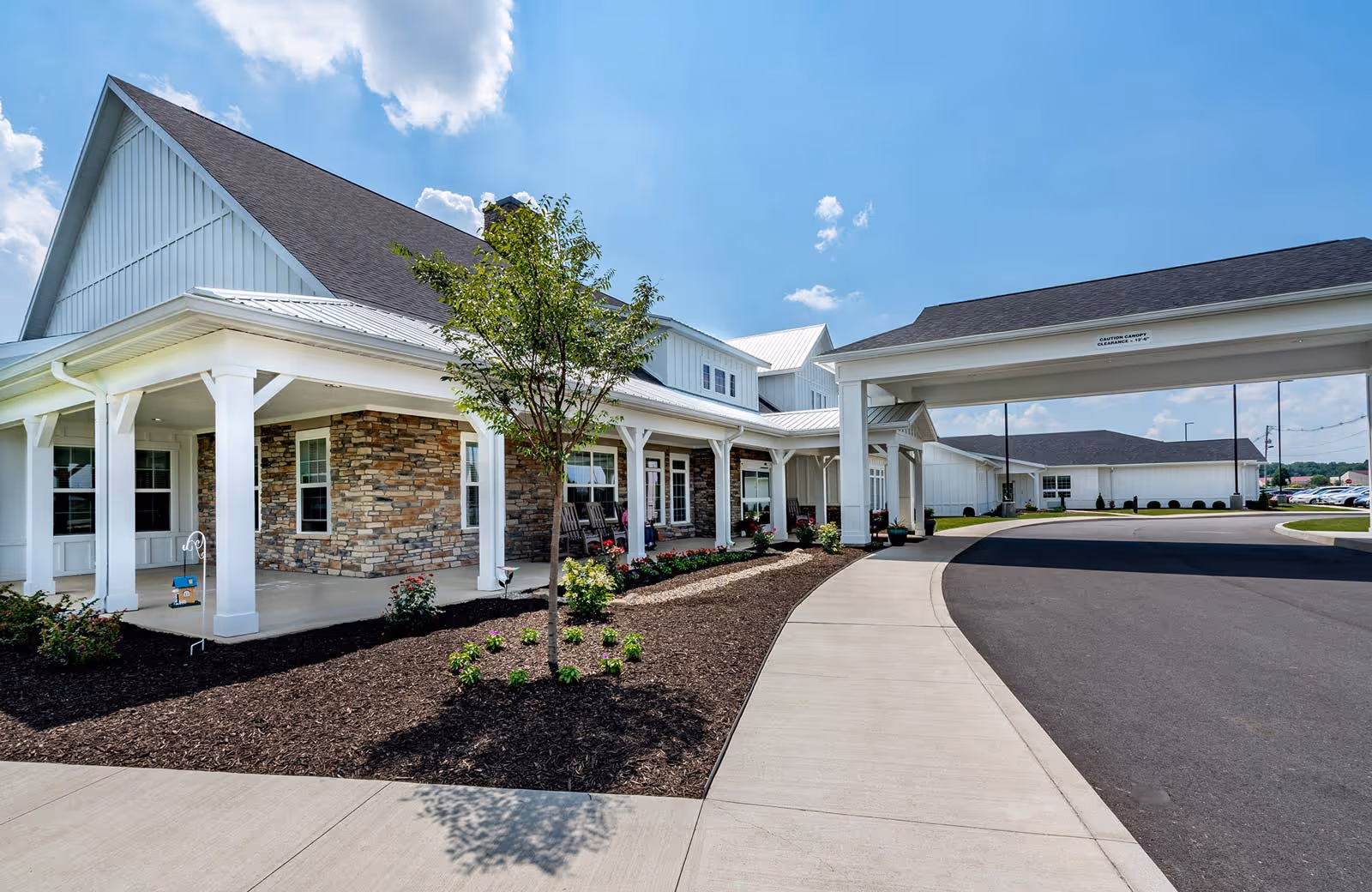 Exterior view of a senior living facility named Sugar Fork Crossing featuring a covered entrance with white pillars, stone and white siding walls, a small landscaped area with a tree and bushes, and a curved driveway under a blue sky with some clouds.