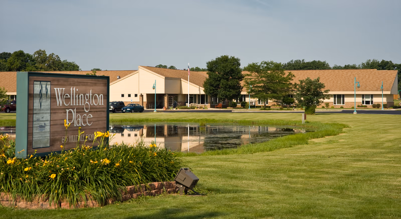 Exterior view of Wellington Place at Hartford, showing a single-story building with a tan roof and beige walls, a pond reflecting the building, green lawn, trees, and a sign with the facility name and address surrounded by yellow flowers.