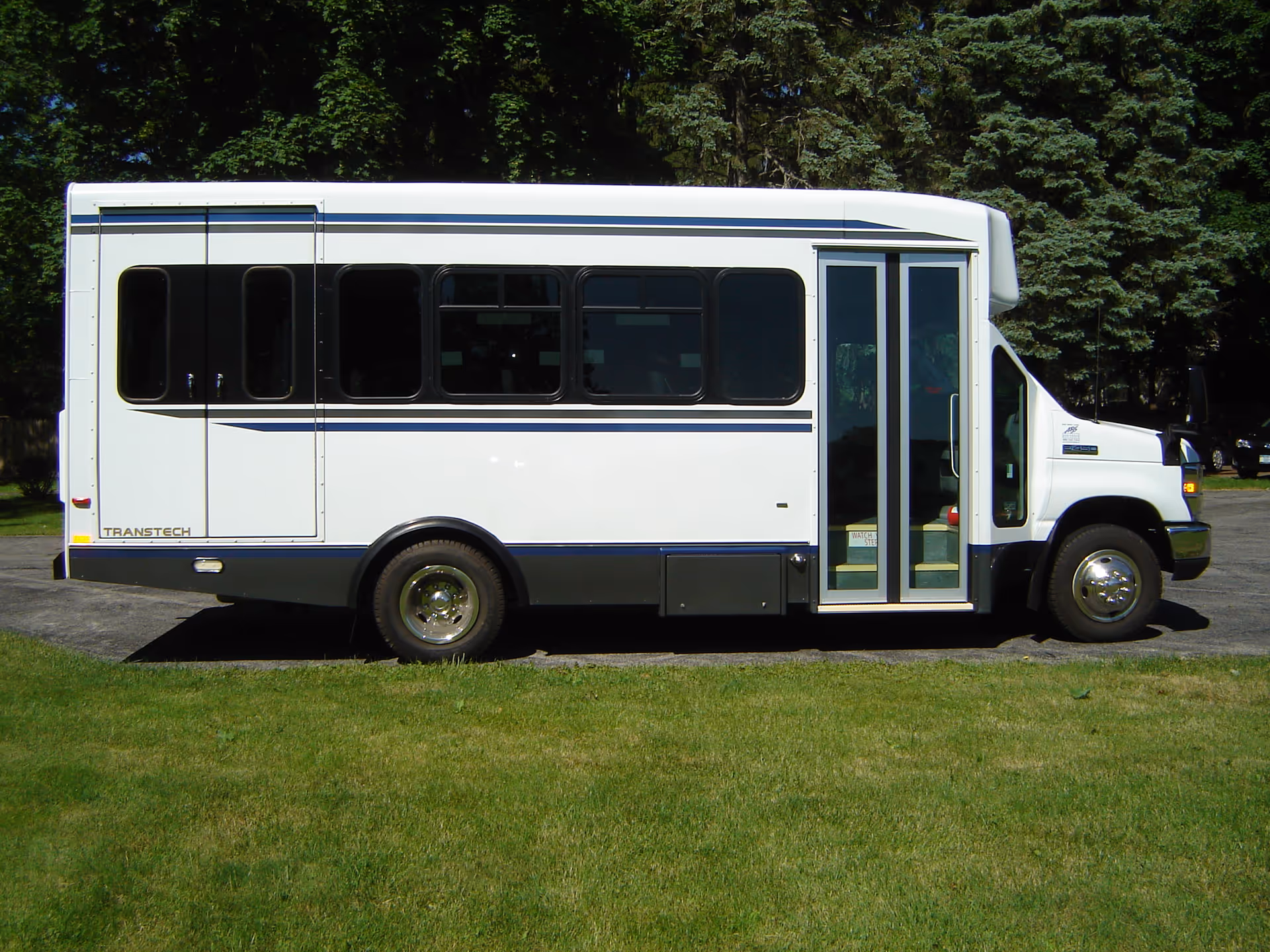 White passenger bus parked on grass with trees in the background.
