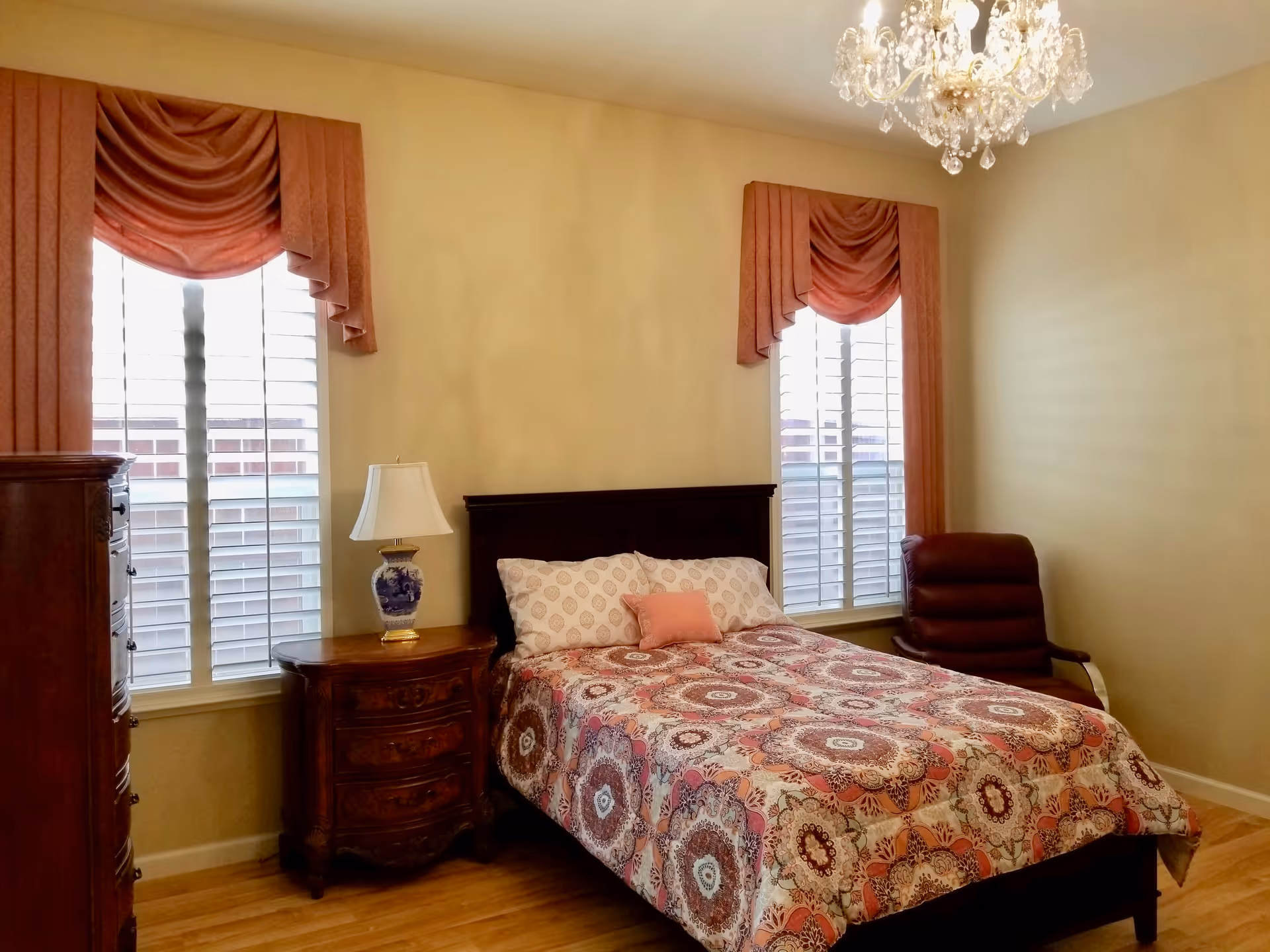 A furnished bedroom with bed, nightstand, dresser, chandelier, and curtains.