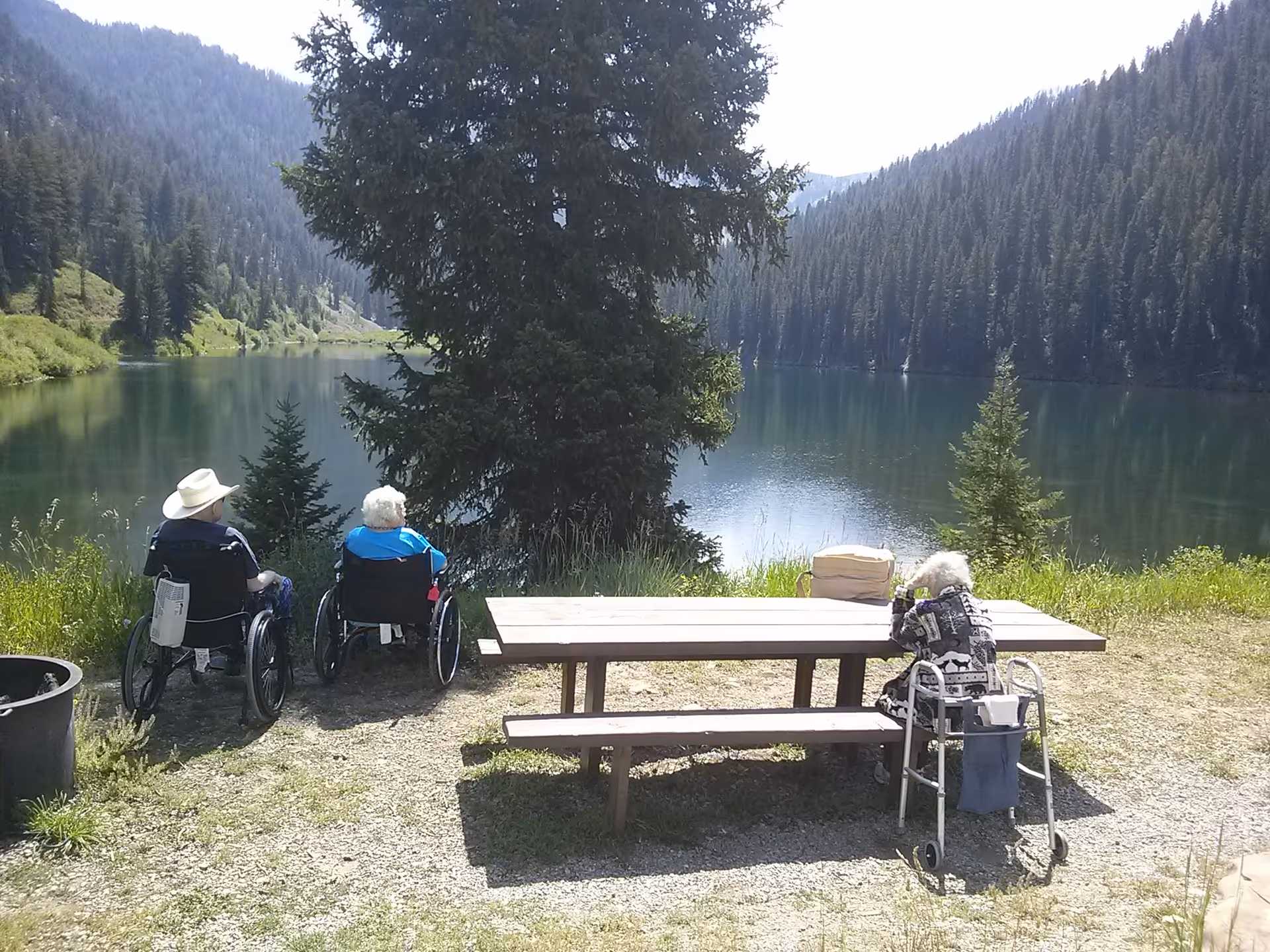Three elderly individuals, two in wheelchairs and one using a walker, are seated outdoors near a wooden picnic table facing a serene lake surrounded by forested hills under a clear sky.