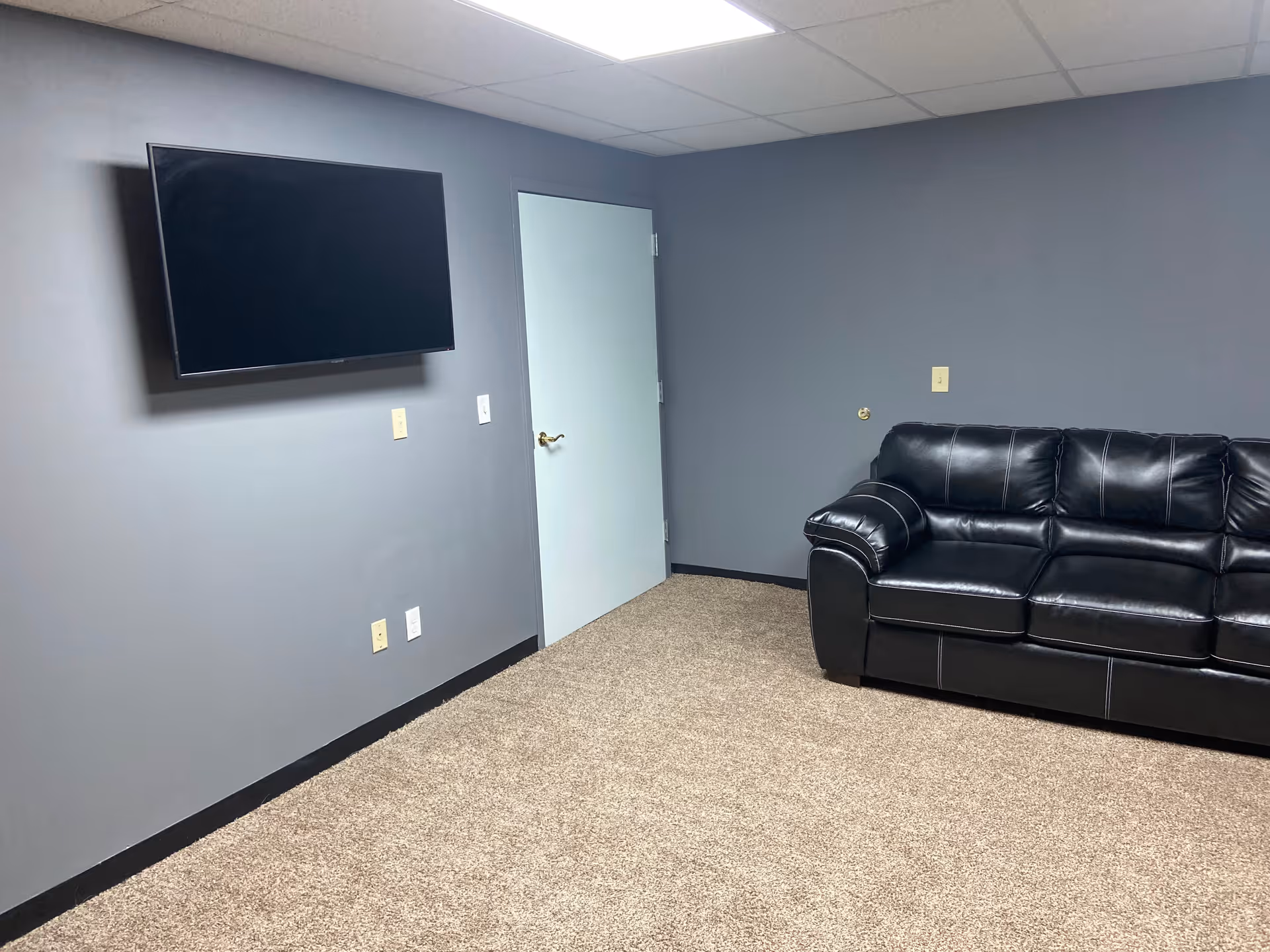 Gray-walled living area with a wall-mounted TV and a black leather sofa.