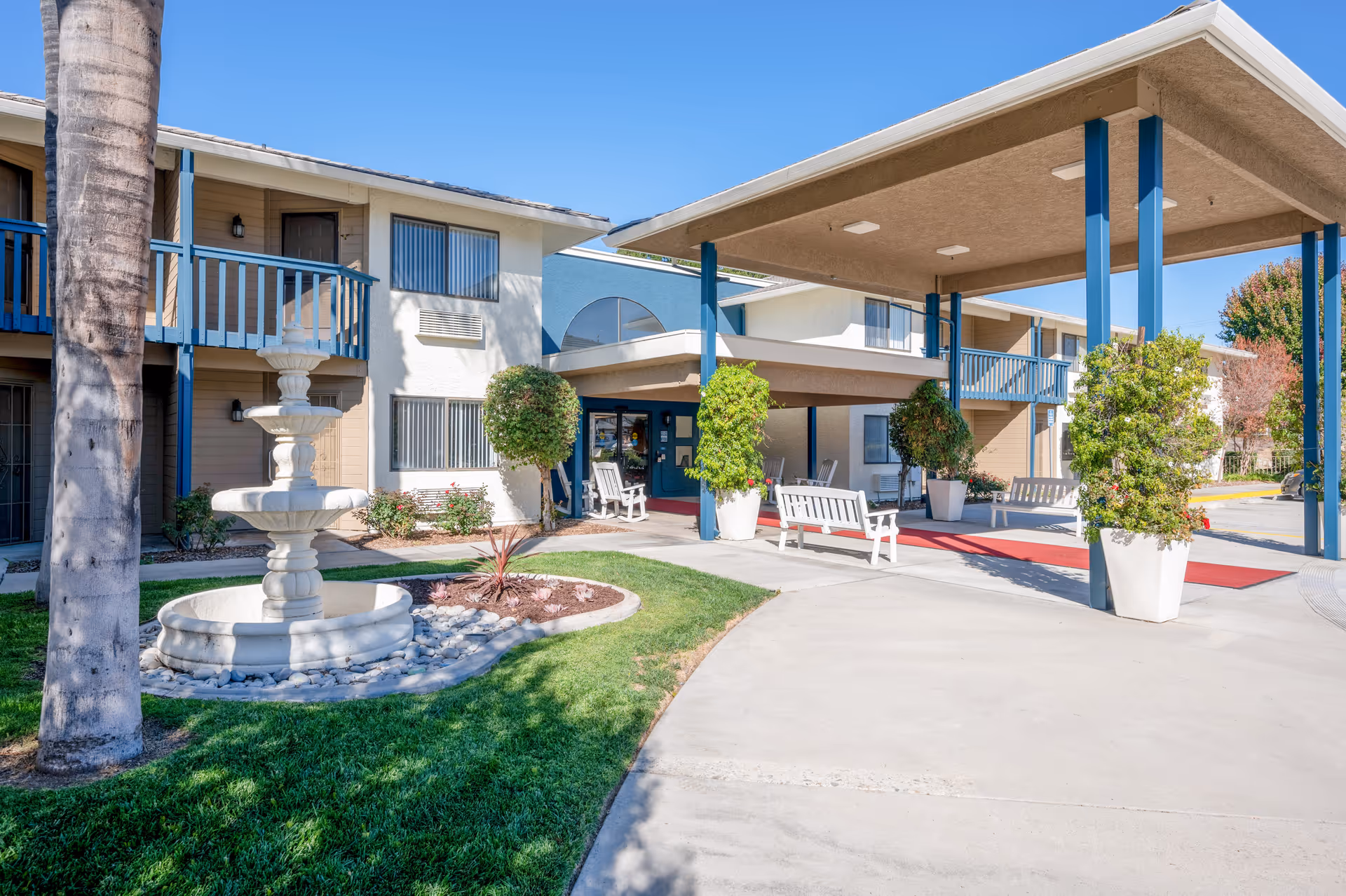 Front entrance of a two-story senior living building with a covered drop-off canopy, benches, potted plants, and a fountain.