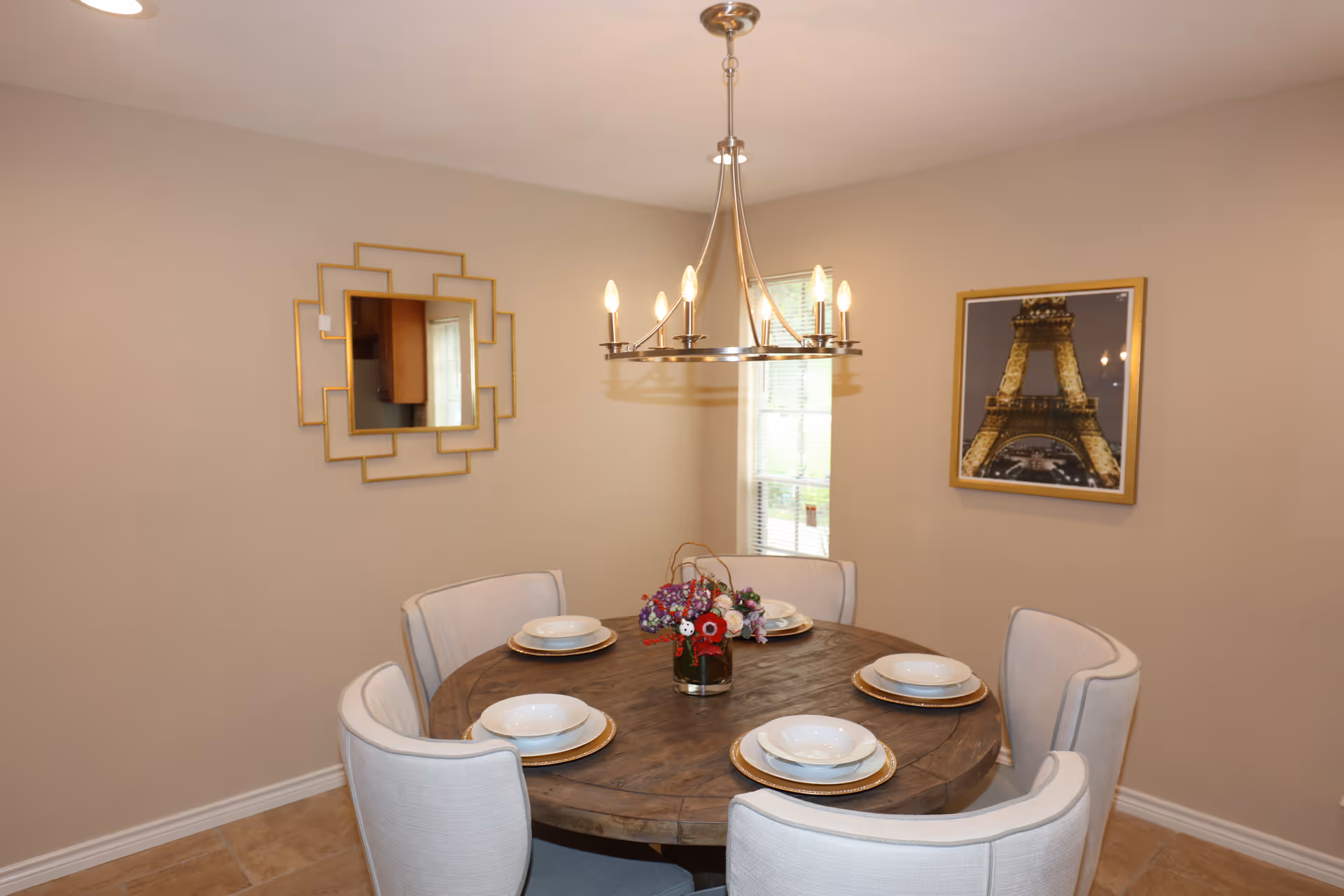 Round wooden dining table with six upholstered chairs, chandelier, and wall art in a dining room.
