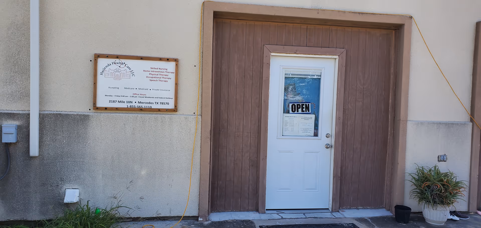 Exterior of Hacienda Health Care facility with a brown door, open sign, wall plaque, and plants.