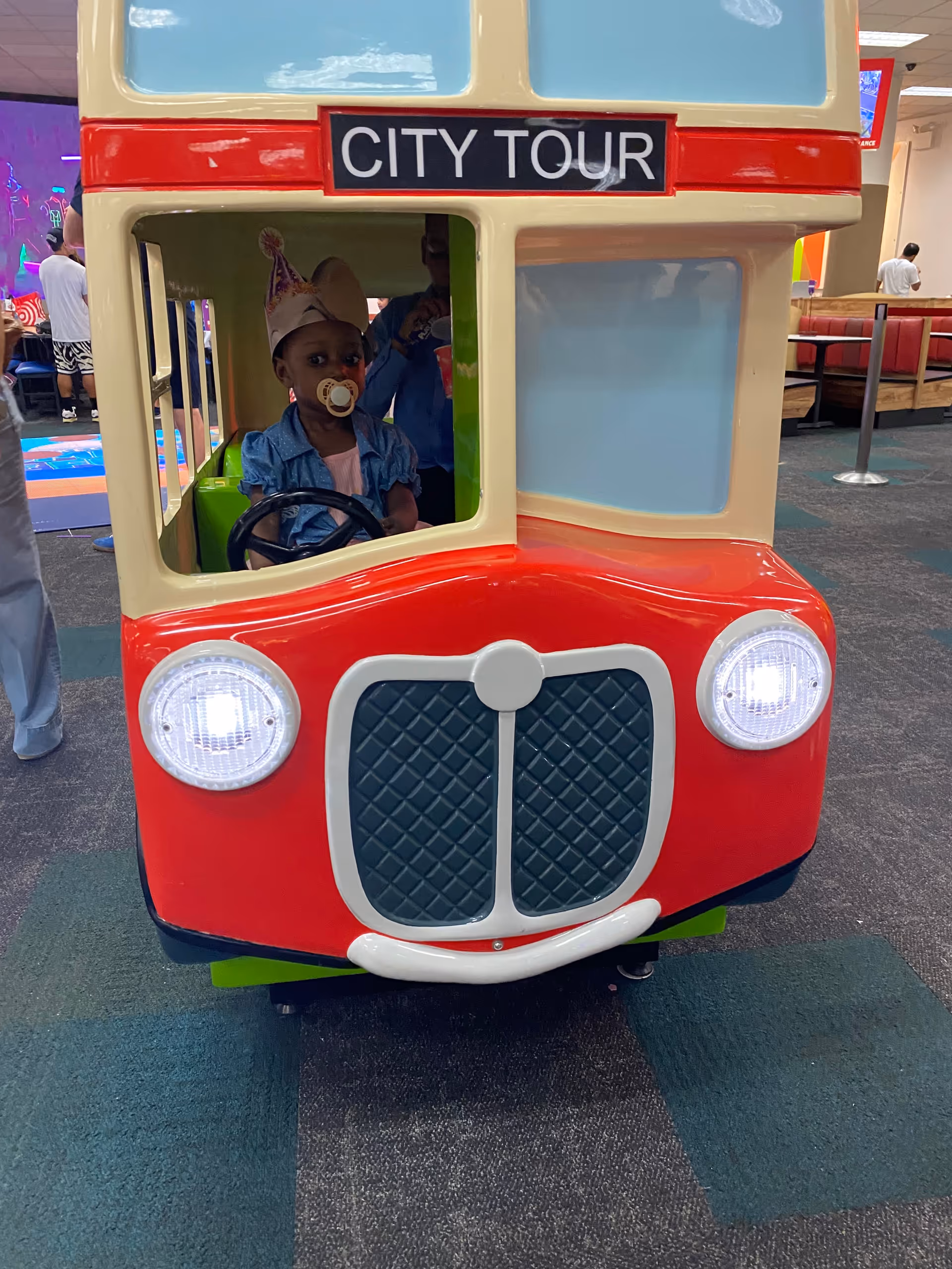 Inside a play area, a red toy bus labeled CITY TOUR with a toddler inside.