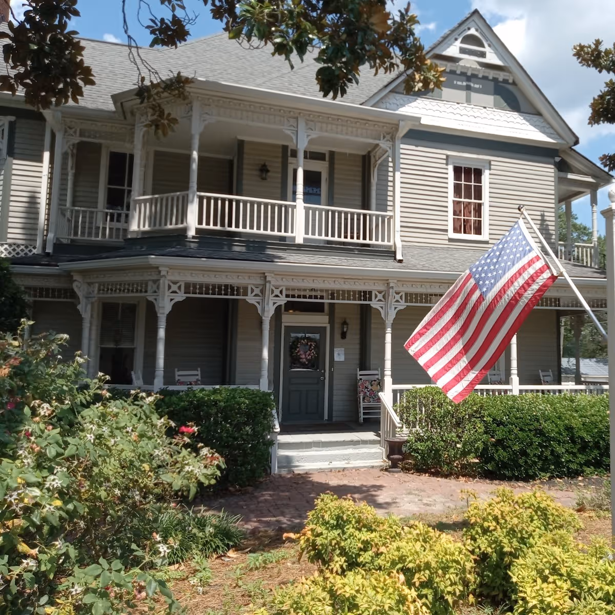 A large Victorian-style house with a wraparound porch, American flag, and landscaped front garden.