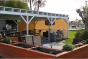 Outdoor covered seating area with chairs and tables, yellow wall backdrop, planters in foreground.