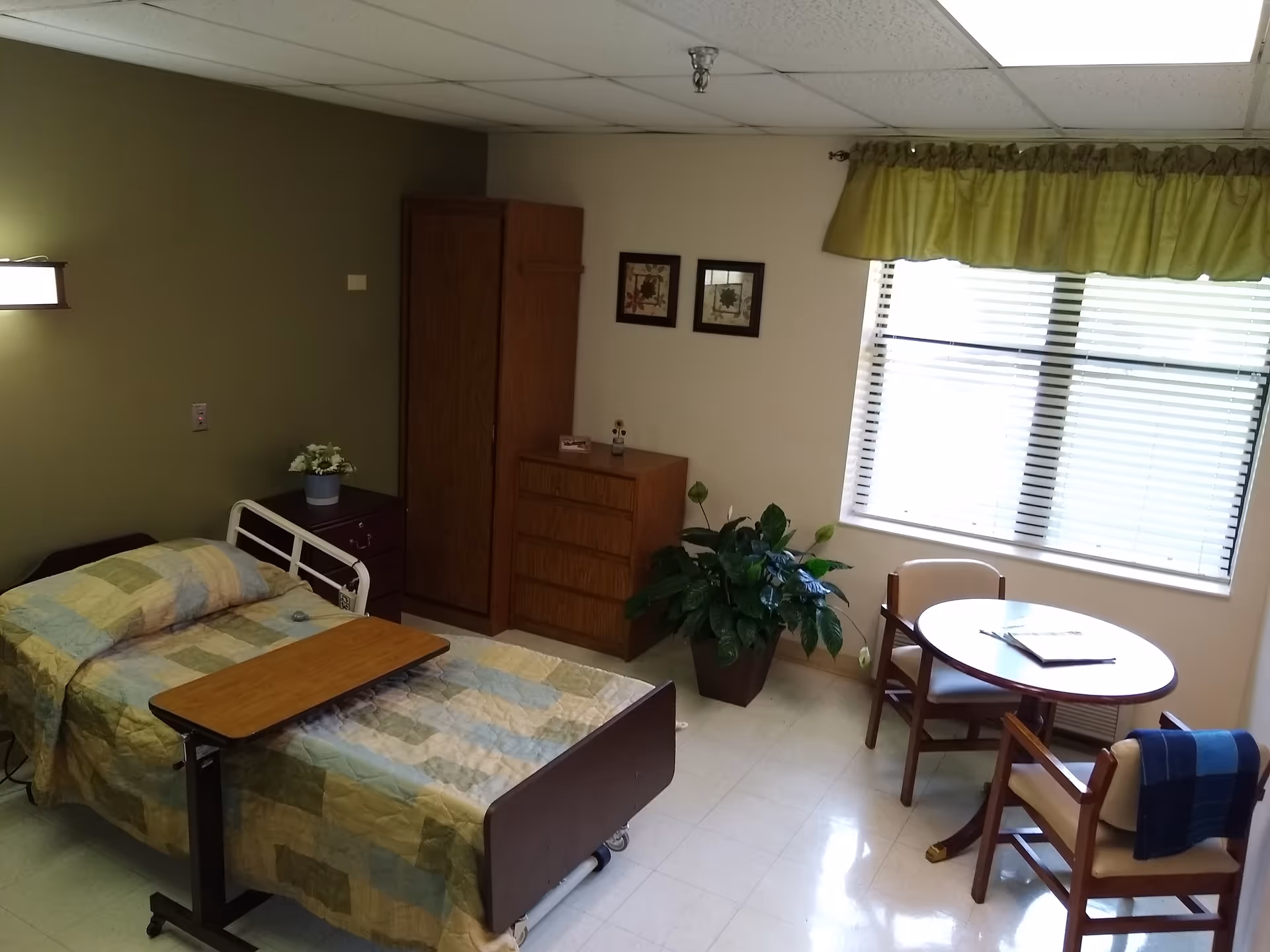 Furnished senior living bedroom with bed, dresser, wardrobe, small table and chairs beside a window.