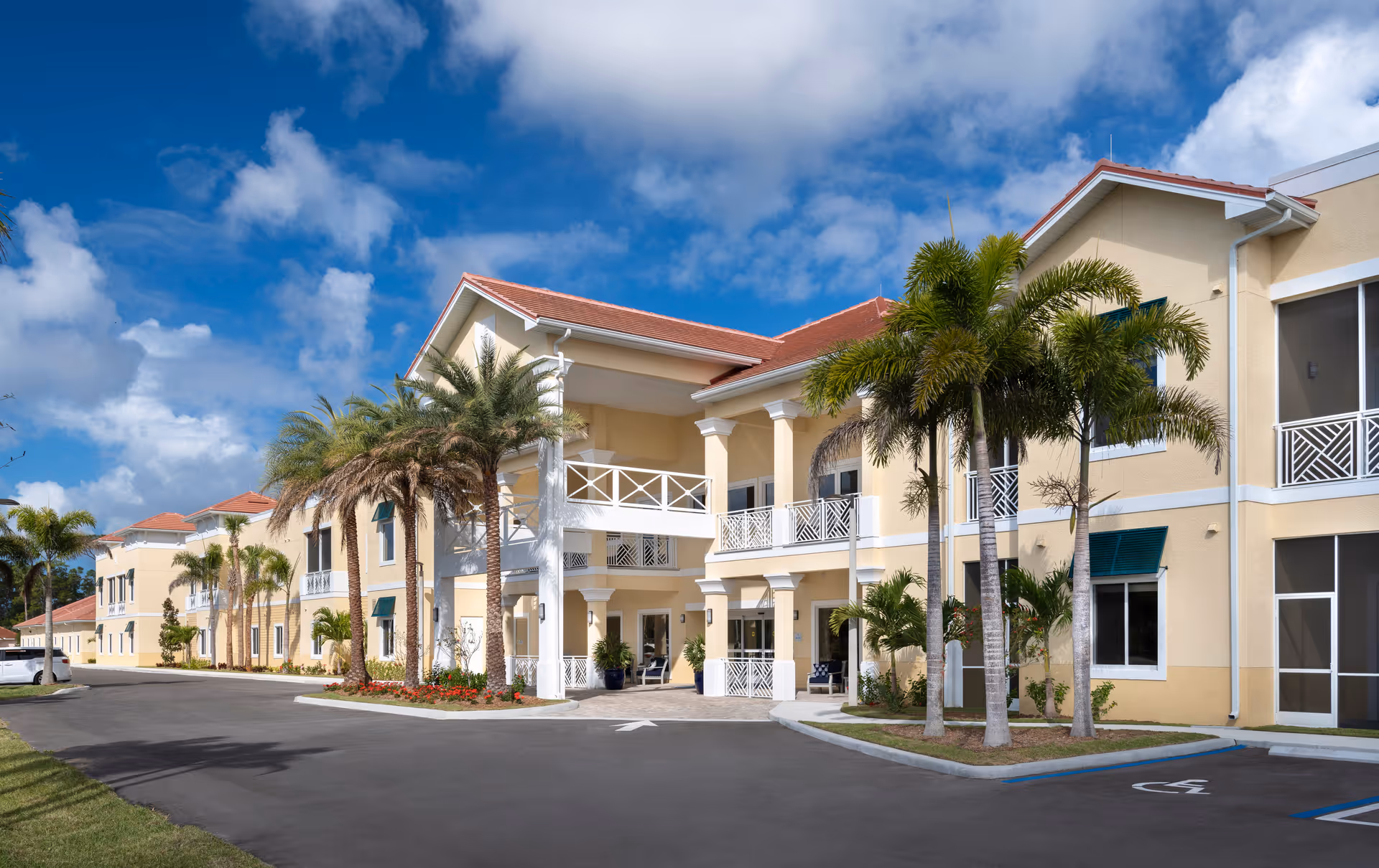 Exterior view of The Pineapple House at Sapphire Lakes, a two-story building with beige walls, white railings, and red-tiled roofs. The entrance is framed by white columns and surrounded by palm trees and landscaped greenery under a partly cloudy blue sky.