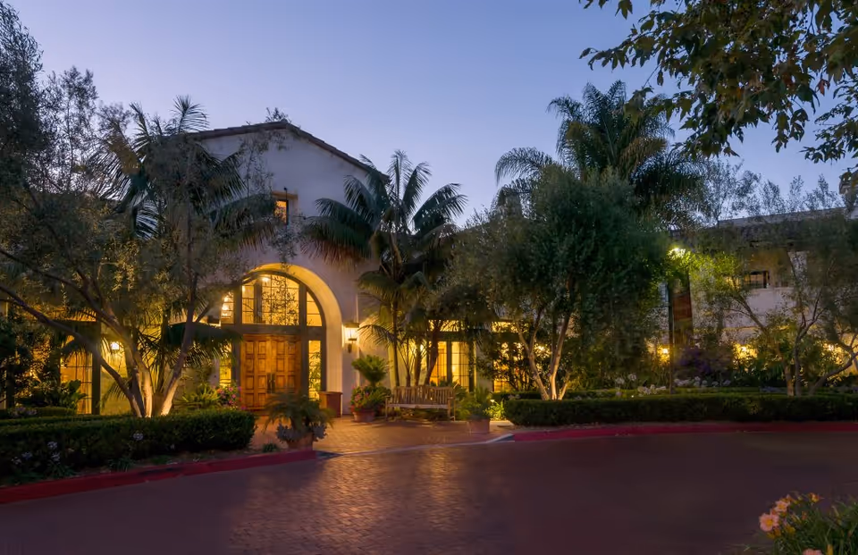 Dusk view of a Mediterranean-style building front with an arched wooden entrance, warm interior lights, palm trees, and a landscaped driveway.