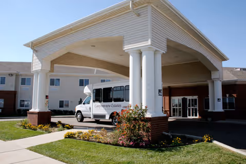 Covered entrance and porte-cochère of a senior living facility with a shuttle van parked under white columns and landscaped lawn.