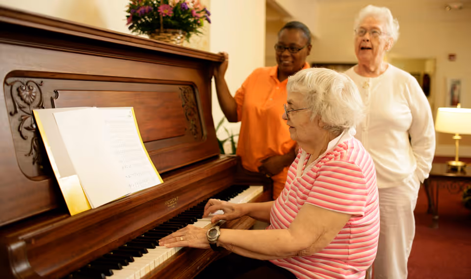 An elderly woman in a pink striped shirt plays a wooden piano with sheet music, while another elderly woman and a caregiver in an orange shirt stand nearby, smiling in a warmly lit room.