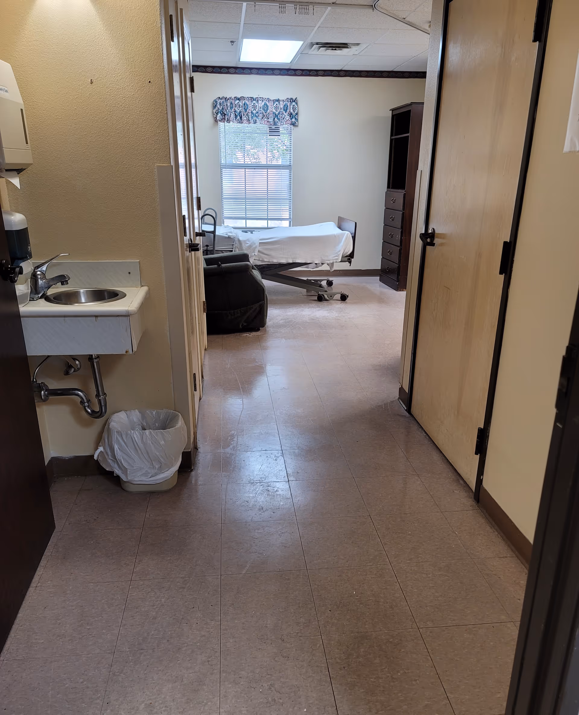 Narrow hallway of a senior living facility opens to patient room with bed, window, and cabinet.