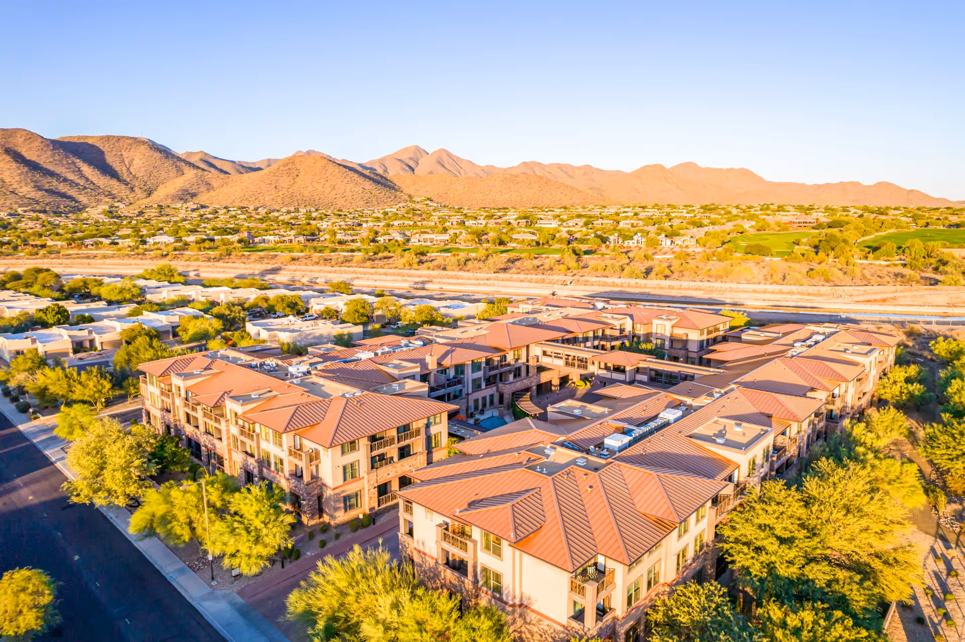Aerial view of a large senior living facility with multiple connected buildings featuring red-tiled roofs, surrounded by trees and desert landscape with mountains in the background under a clear sky.