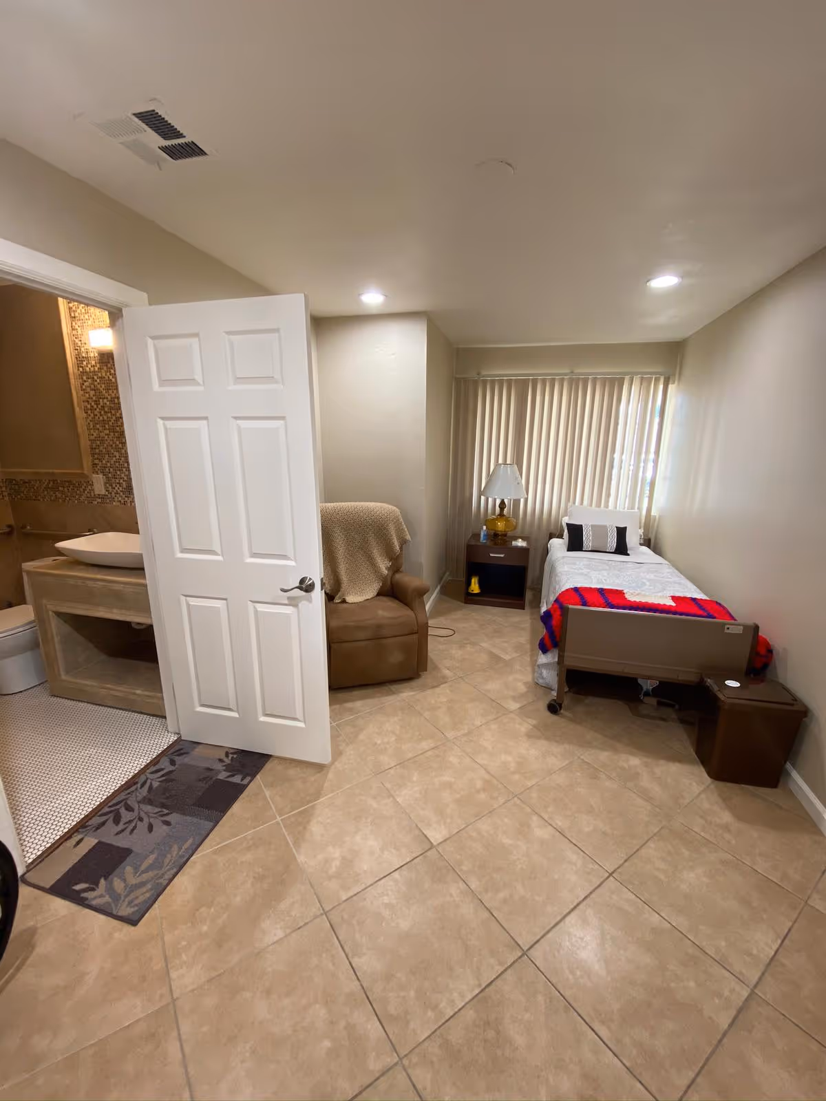 Senior living bedroom with a bed, recliner, nightstand, lamp, and open bathroom doorway.