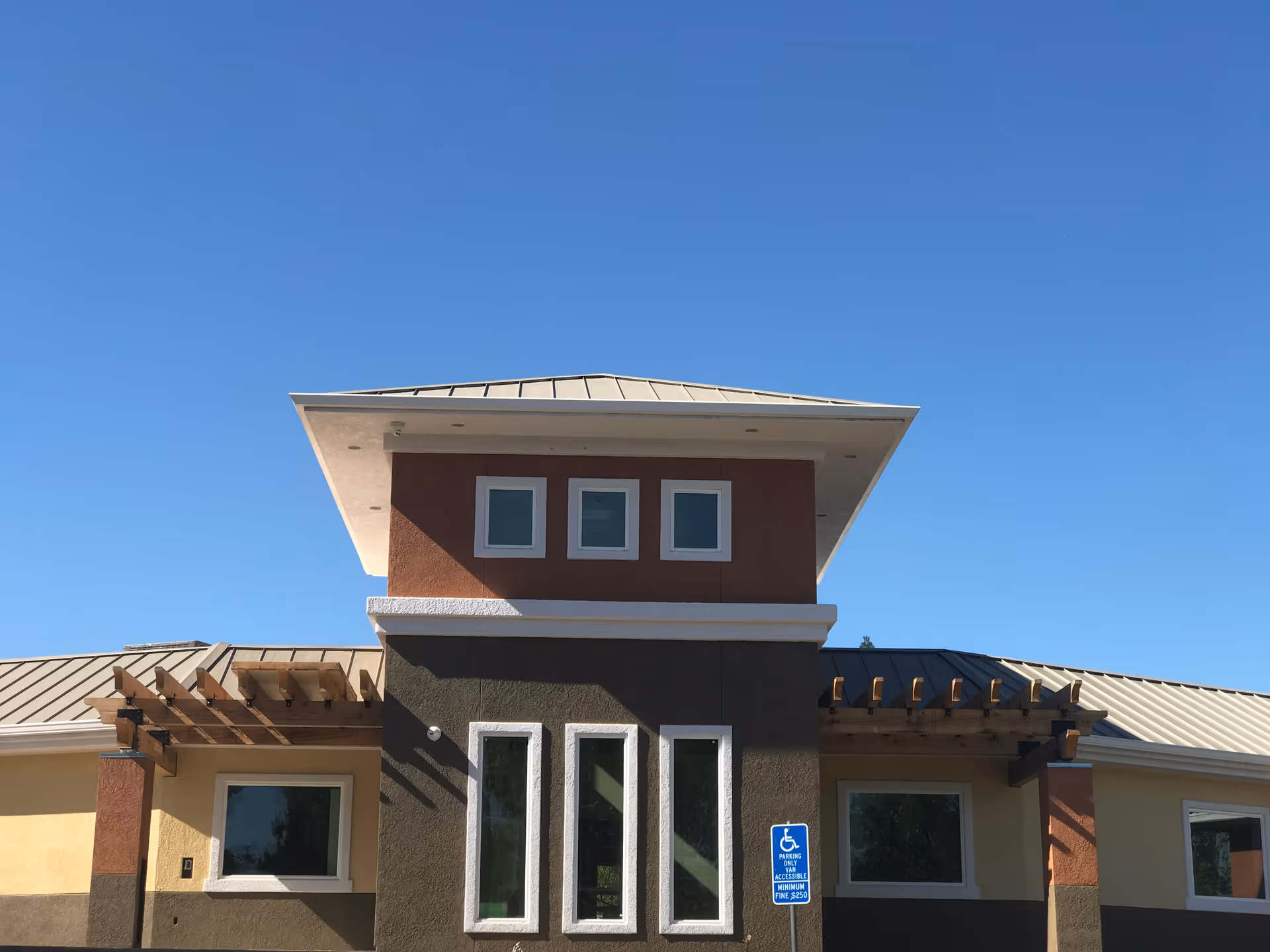 Exterior front view of a modern senior living facility building with a beige, brown, and dark green color scheme, featuring three small square windows near the roof, two larger vertical windows below, and a metal roof under a clear blue sky. There is a handicap parking sign visible in front of the building.