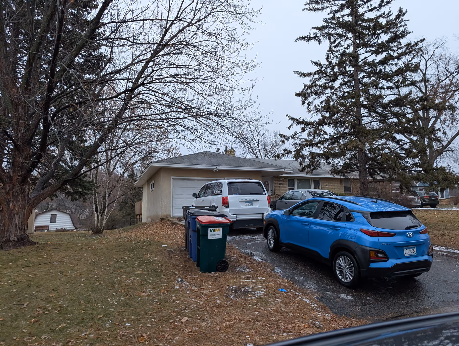 Single-story house front with a driveway, several parked cars and trash bins beneath leafless trees on an overcast day.